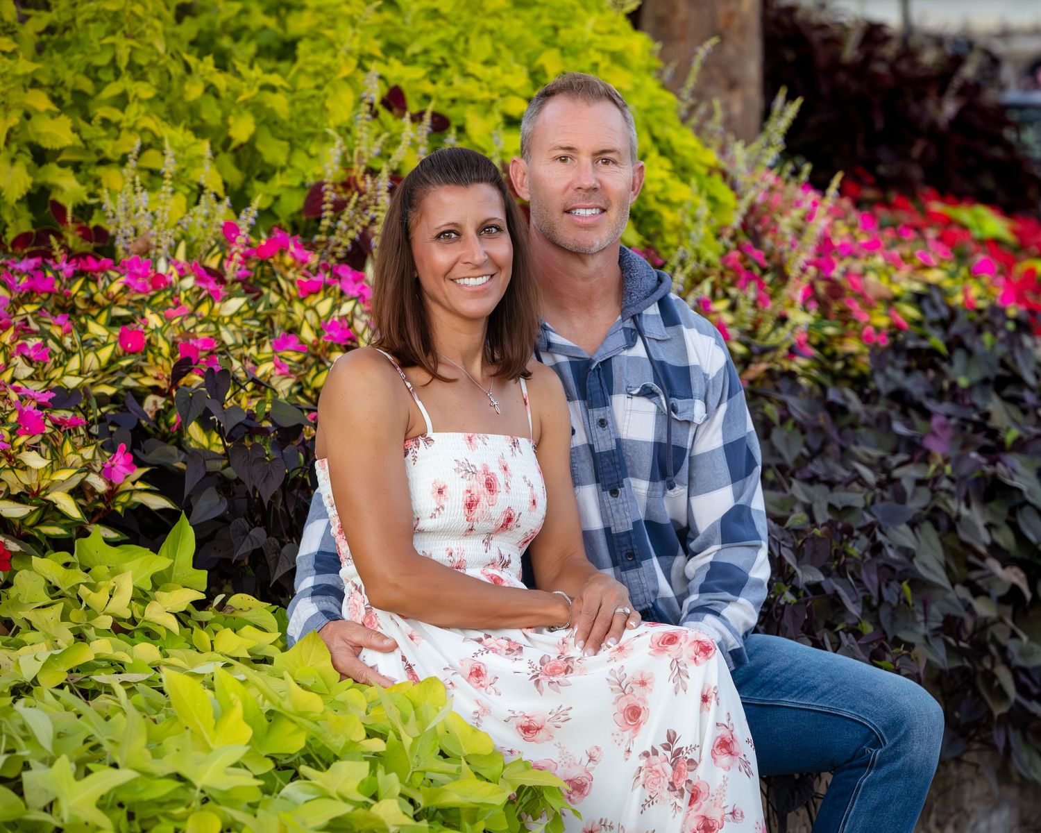 Smiling couple sitting together outdoors surrounded by vibrant pink flowers and lush green foliage in a garden setting.