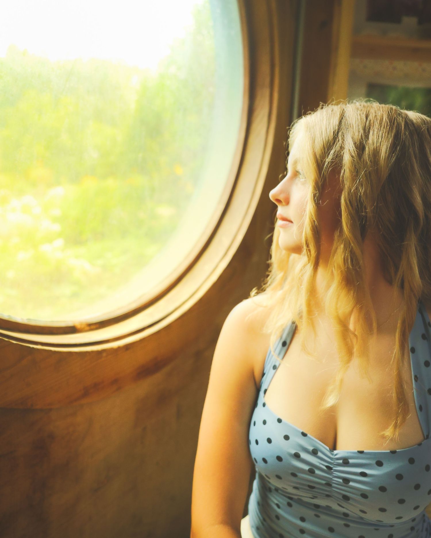 Young woman in polka dot dress gazing through a round porthole window with sunlit greenery outside.