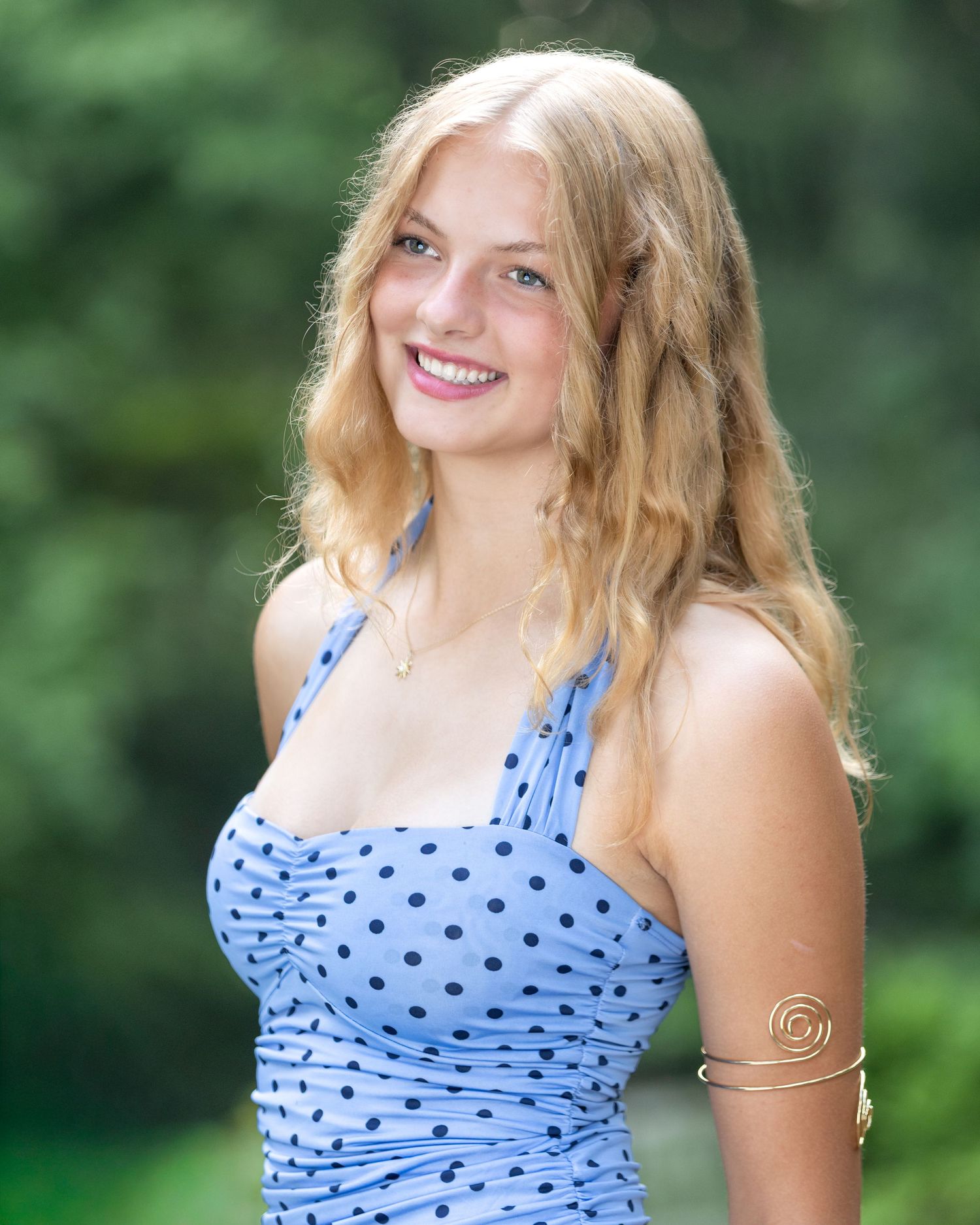 Smiling young woman with blonde hair wearing a blue polka dot halter dress outdoors with green foliage background.