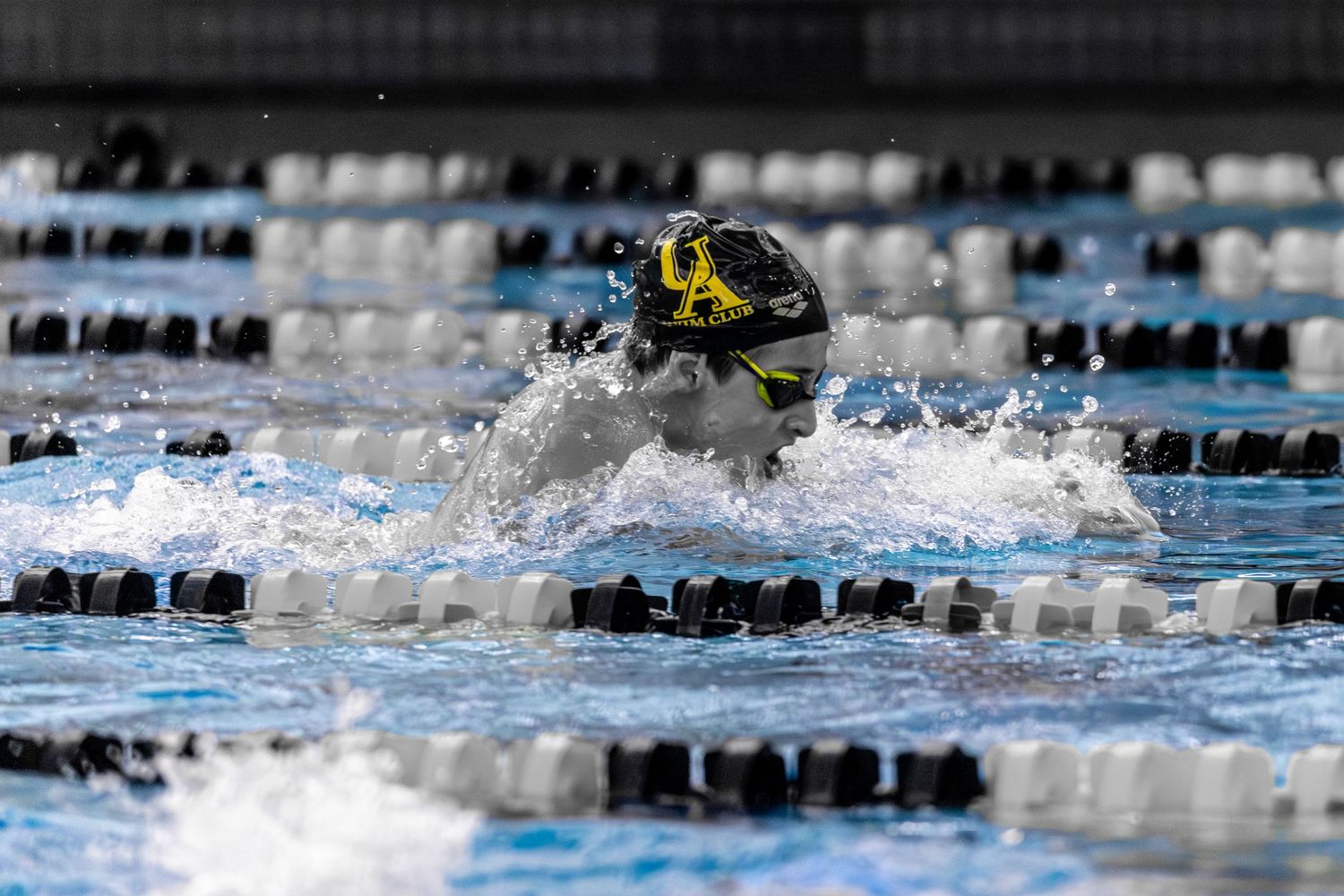 Competitive swimmer performing breaststroke in a pool during a swim meet, wearing a black cap and goggles.