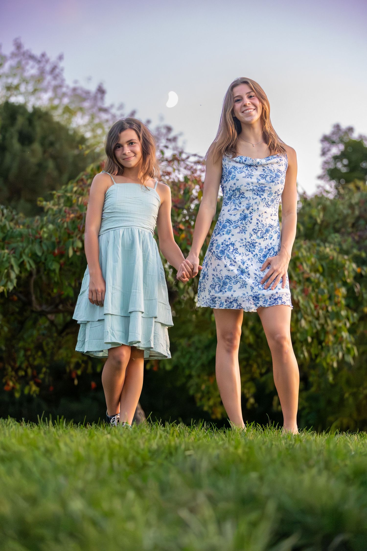 Two young girls in summer dresses holding hands on green grass with trees and moon in background.