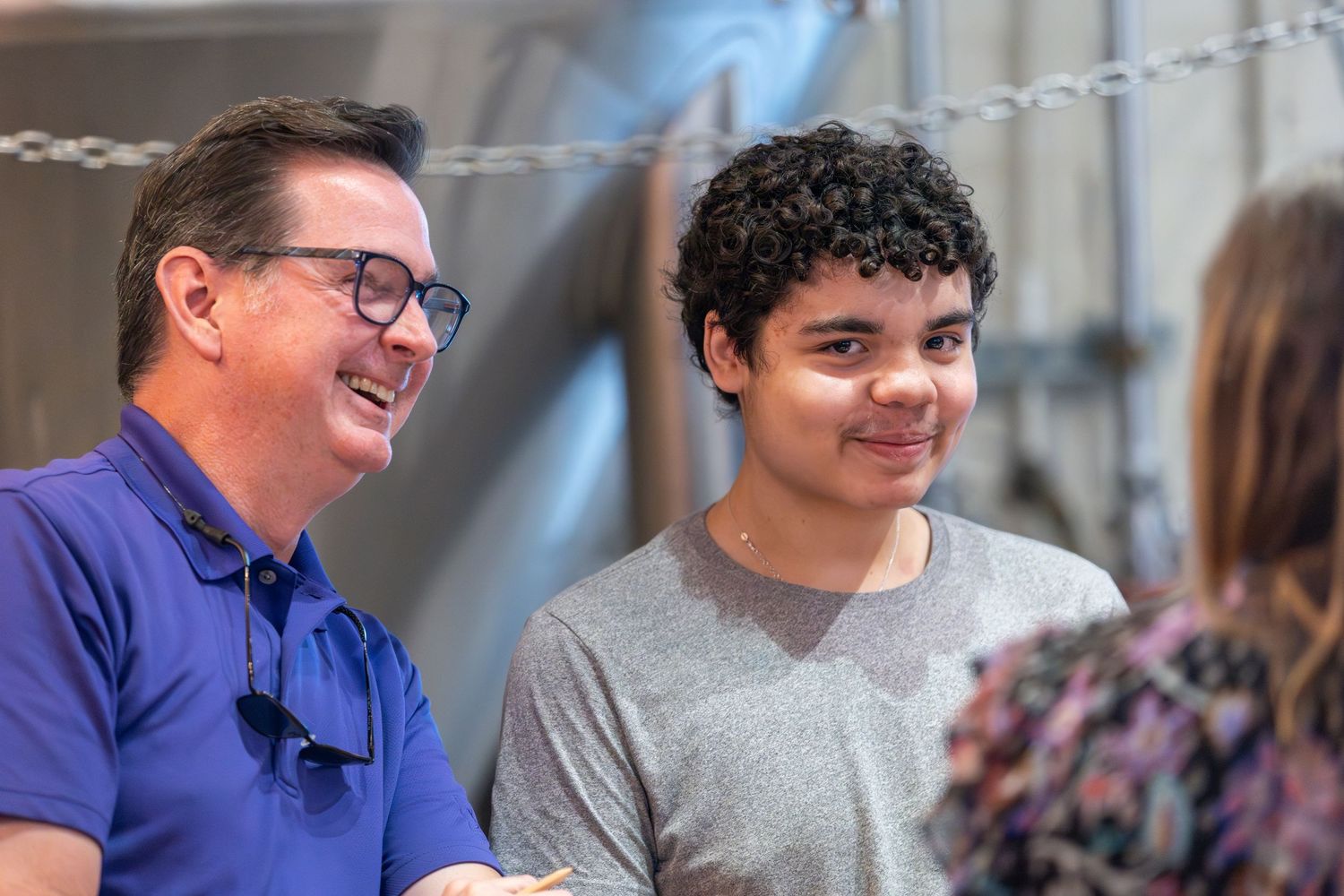 A smiling man in a blue polo shirt sits beside a teenage boy in a gray shirt, both engaged in cheerful conversation indoors.