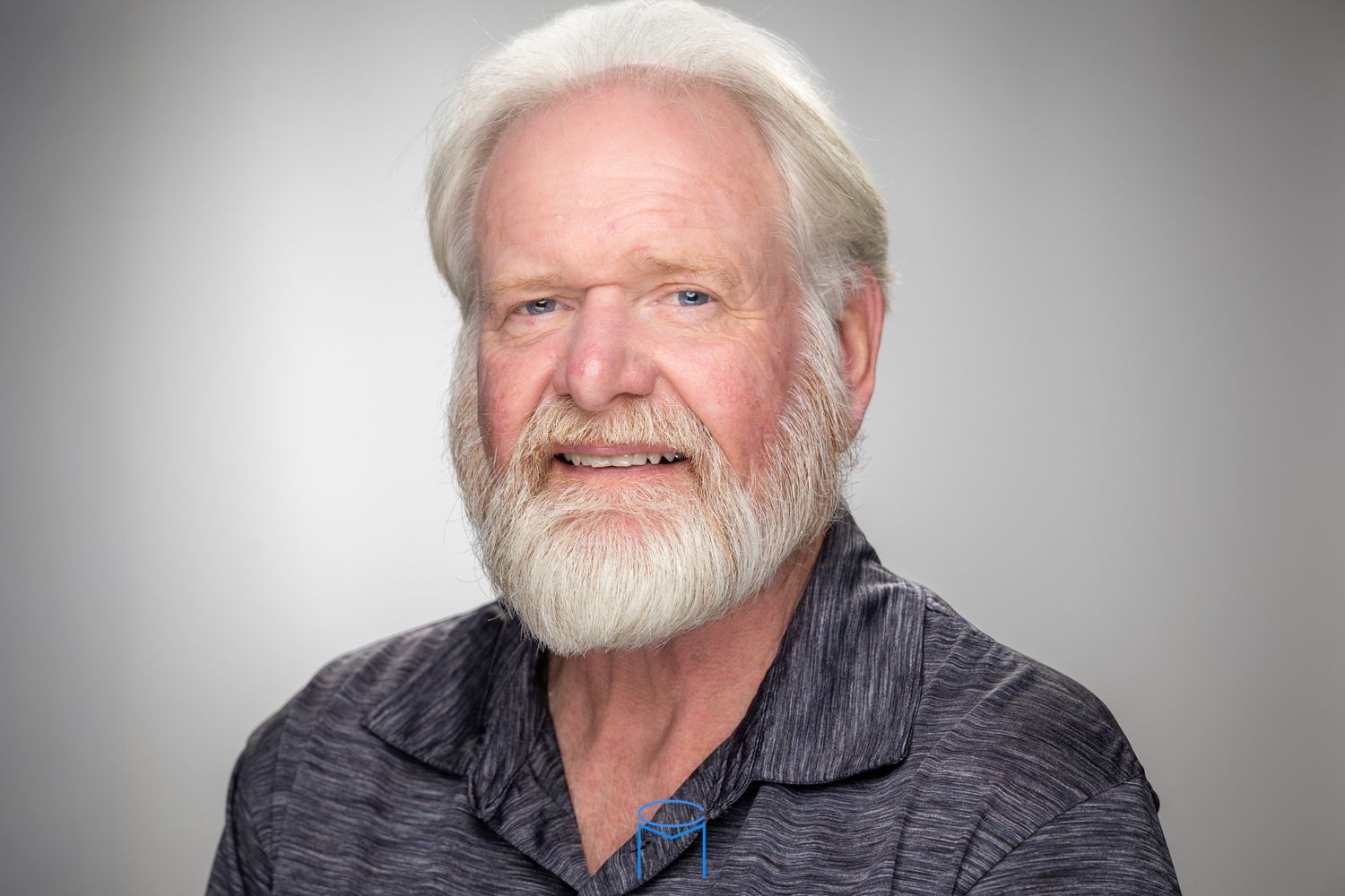 Smiling elderly man with white hair and beard wearing a gray shirt against a neutral background.