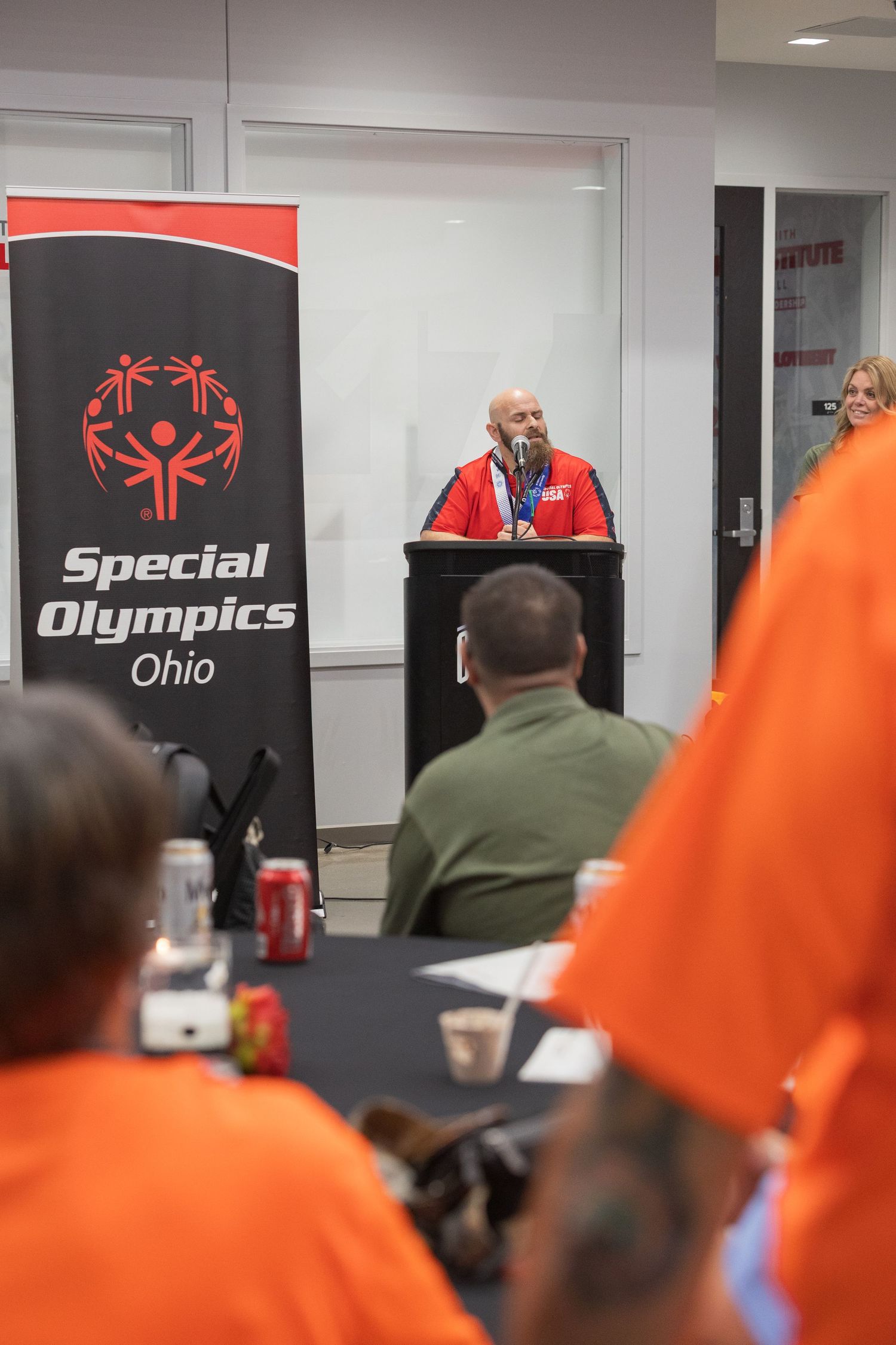 Man speaking at podium beside Special Olympics Ohio banner to audience in orange shirts.