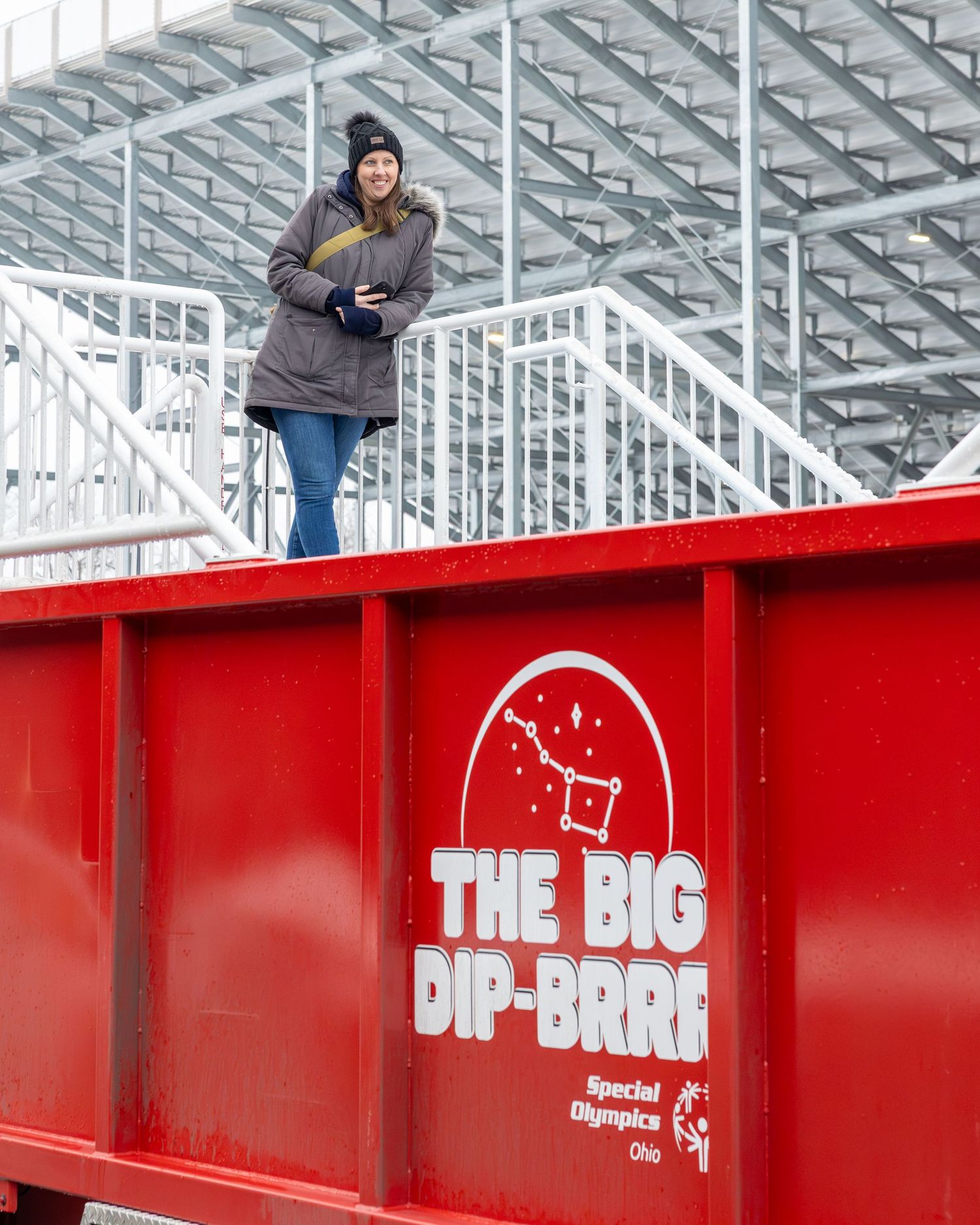 Woman standing on a red Special Olympics Ohio The Big Dip-Brrr dumpster wearing winter coat and hat outdoors.