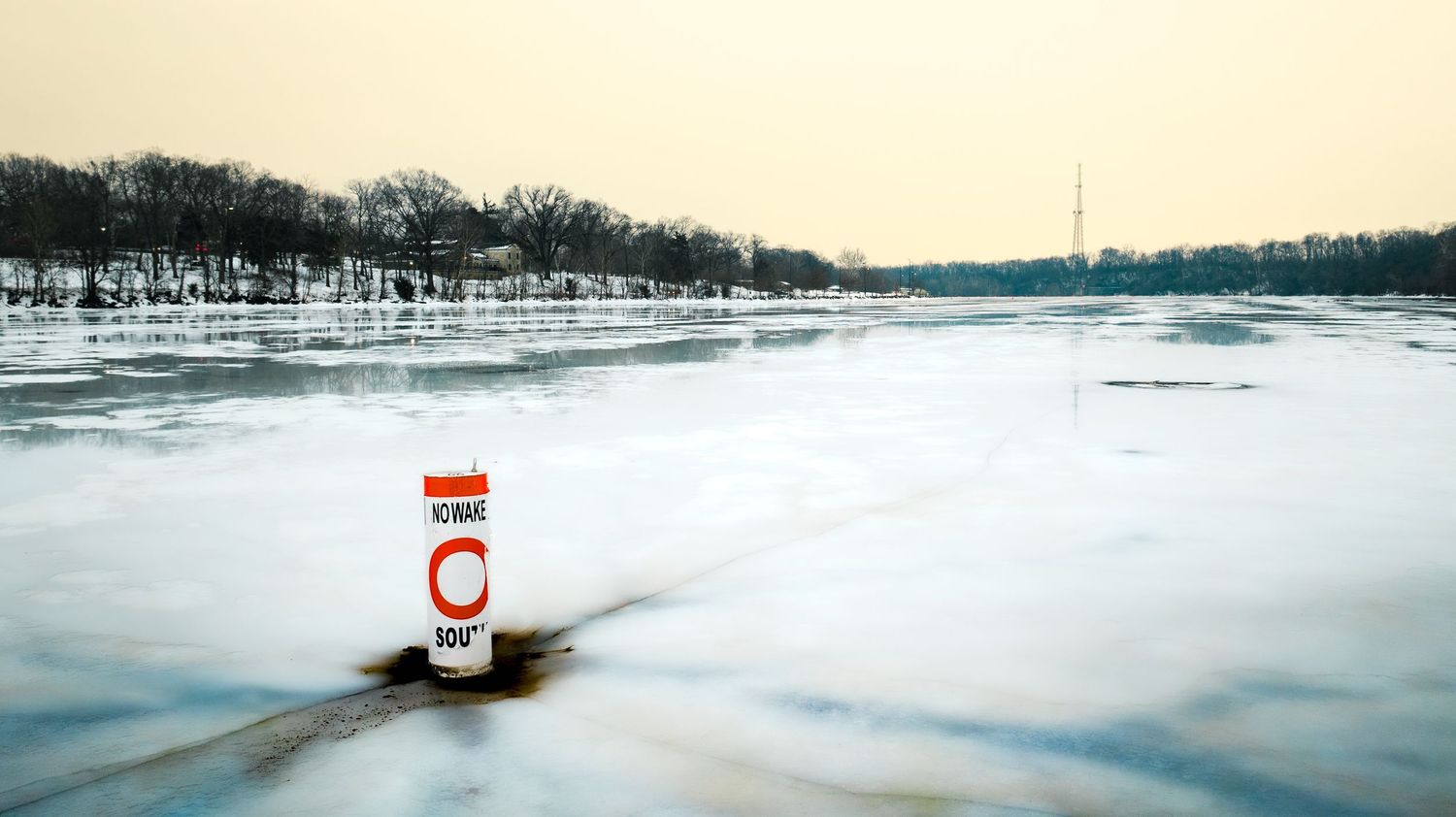 A red and white channel marker stands in a frozen river surrounded by ice and snow-covered trees in winter.
