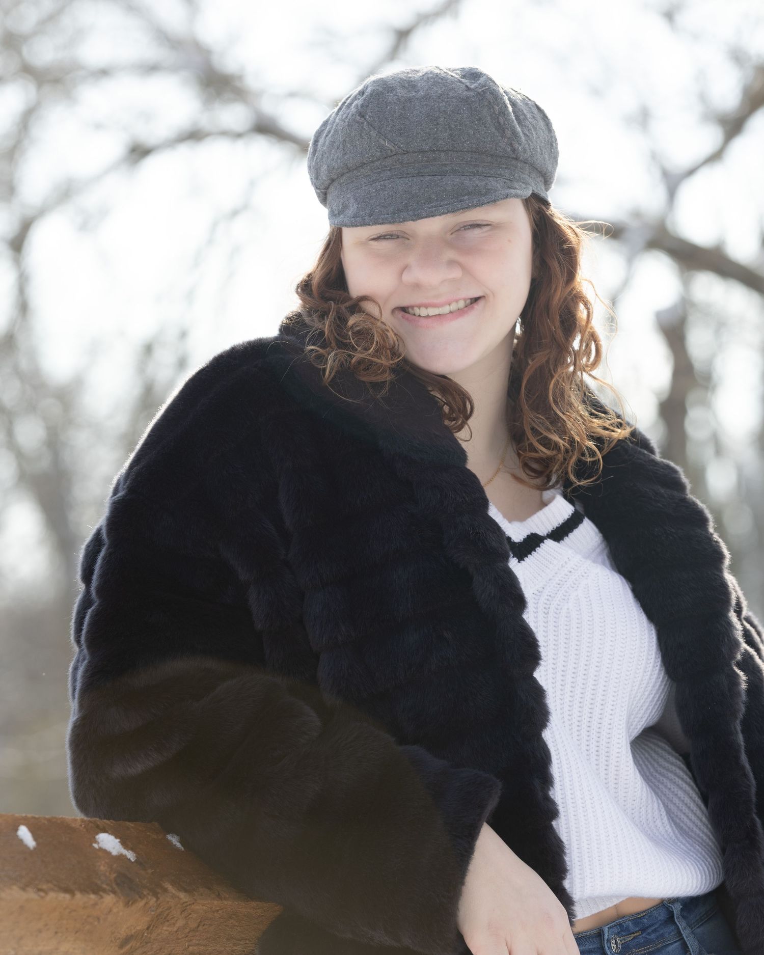 Young woman smiling outdoors in winter, wearing black fur coat, white top, and gray newsboy cap with curly auburn hair.