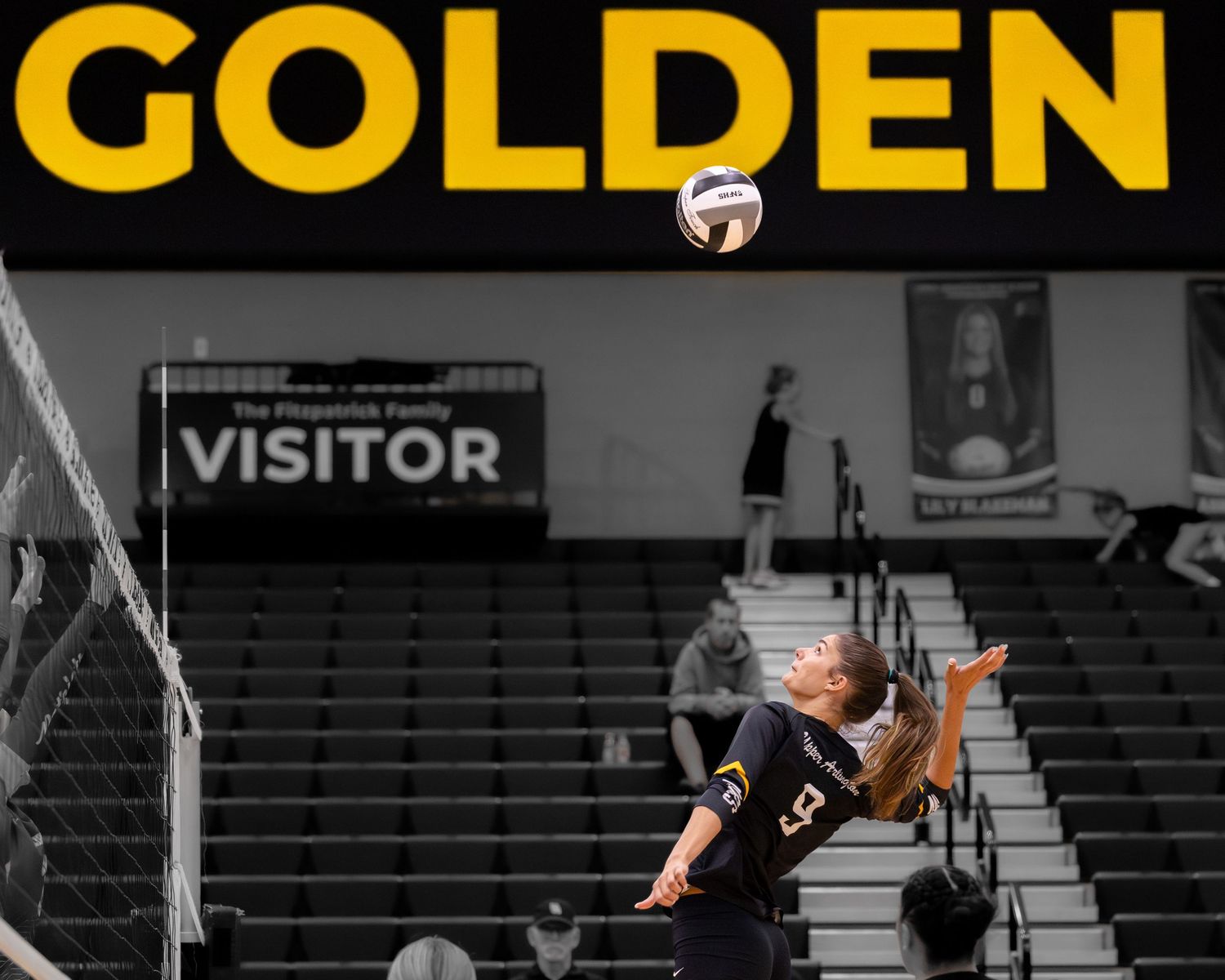 Volleyball player serving the ball during a match, with GOLDEN text overlay in black and gold colors.
