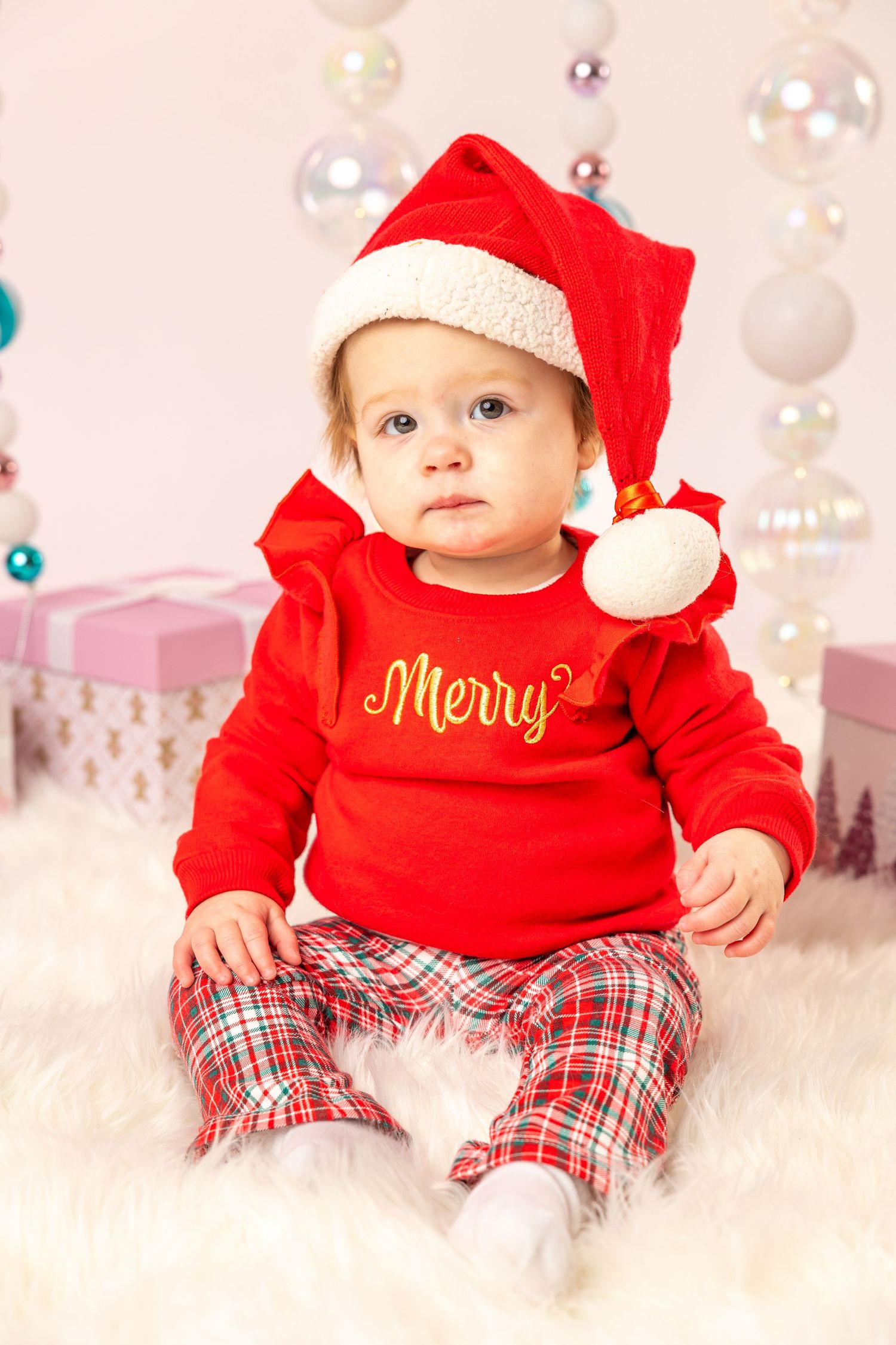 Adorable baby in red Merry sweater, Santa hat, and plaid pants sitting on fur rug with Christmas ornaments.