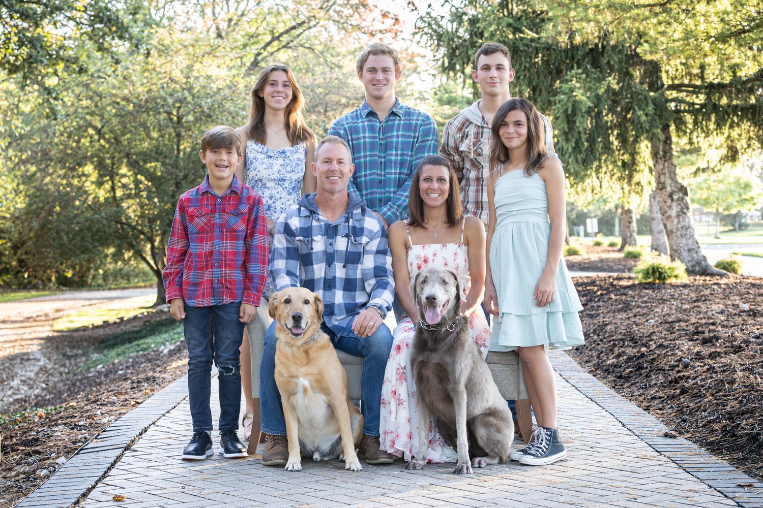 A group poses together with a golden retriever dog on a sunlit outdoor path surrounded by trees and fallen leaves.