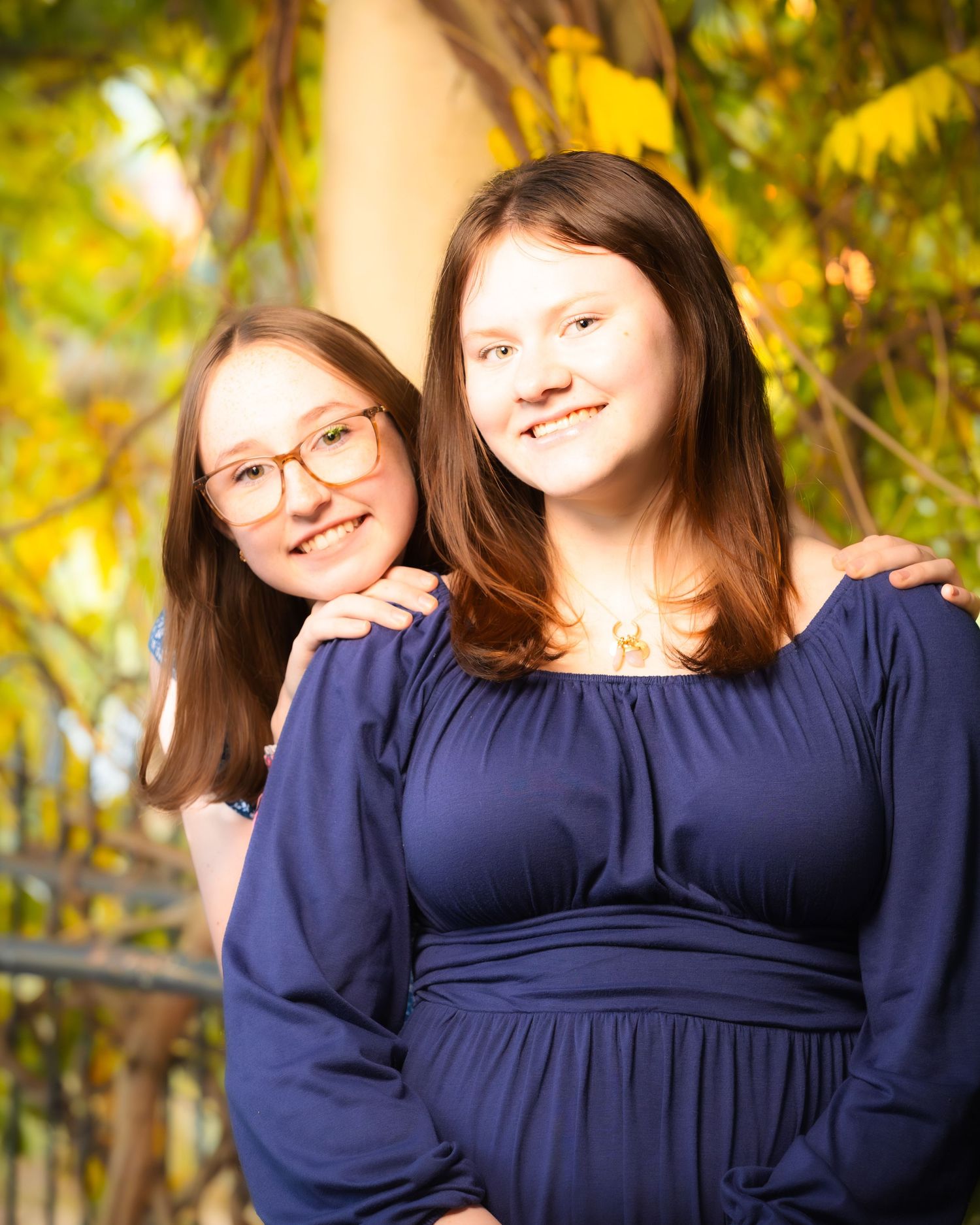 Two young women in matching navy blue dresses smile together outdoors with golden autumn foliage in the background.