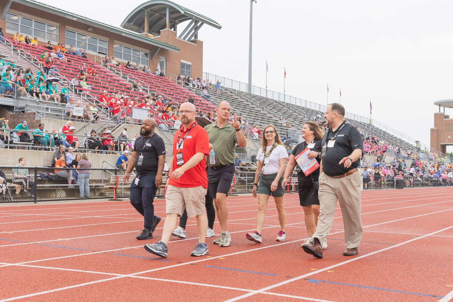 Group of people walking on a red track at a large stadium filled with spectators during an outdoor sporting event.