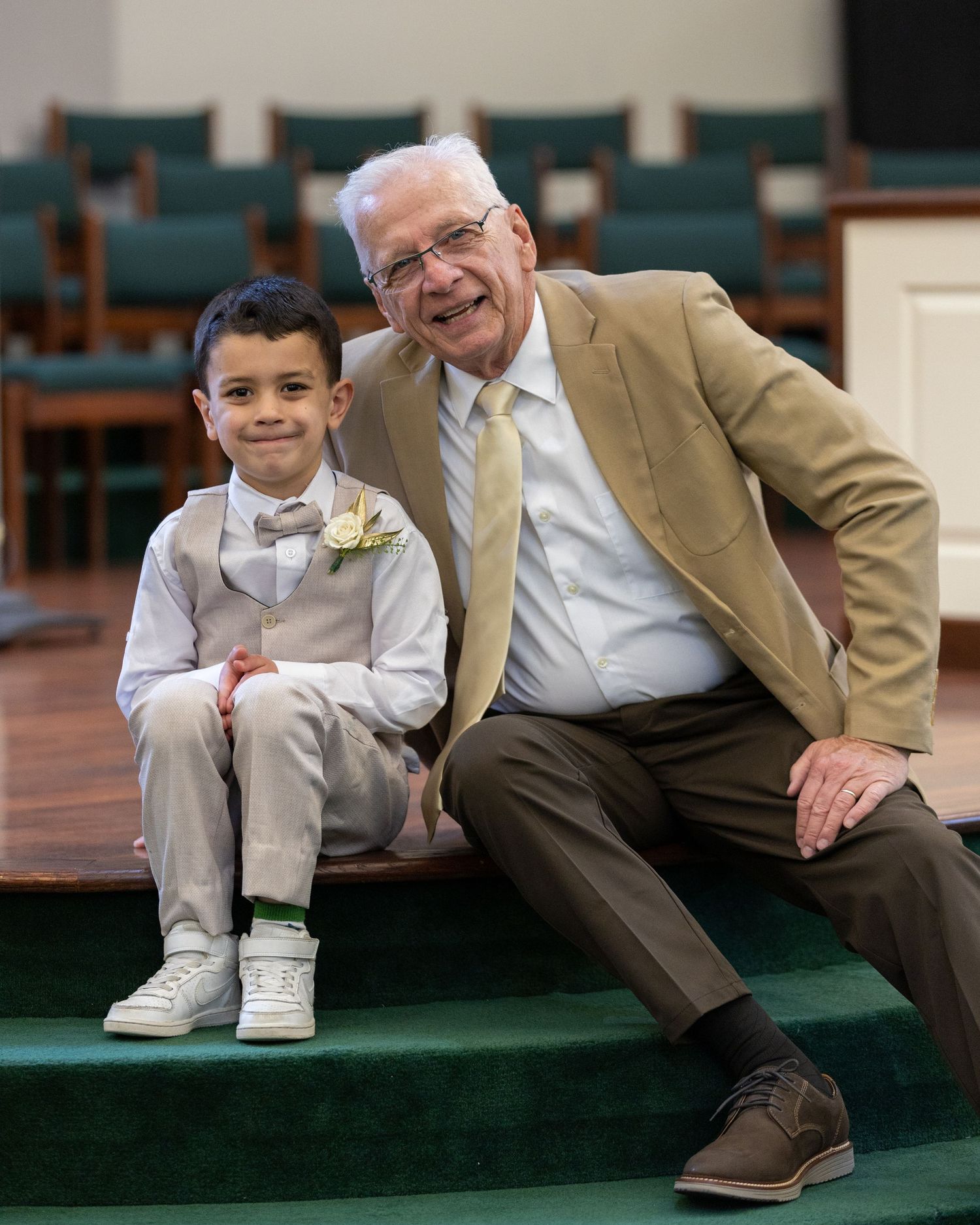 An elderly man and young boy in formal attire smile together on church steps at a wedding ceremony.