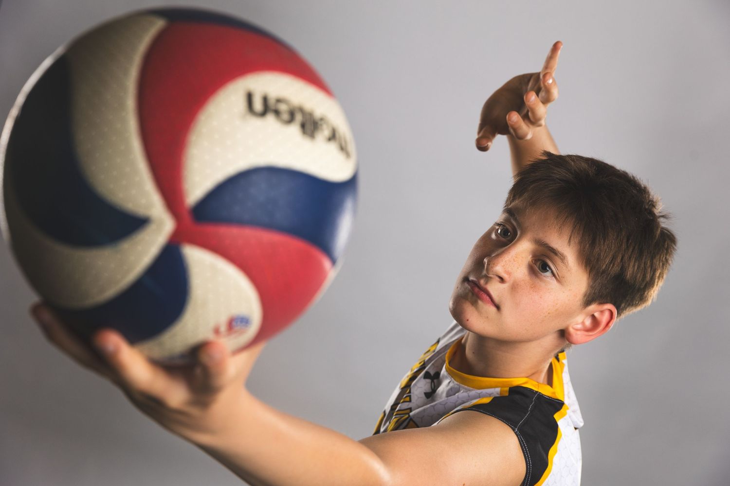 Young boy in volleyball jersey holding a Molten volleyball, pointing upward against a gray background.