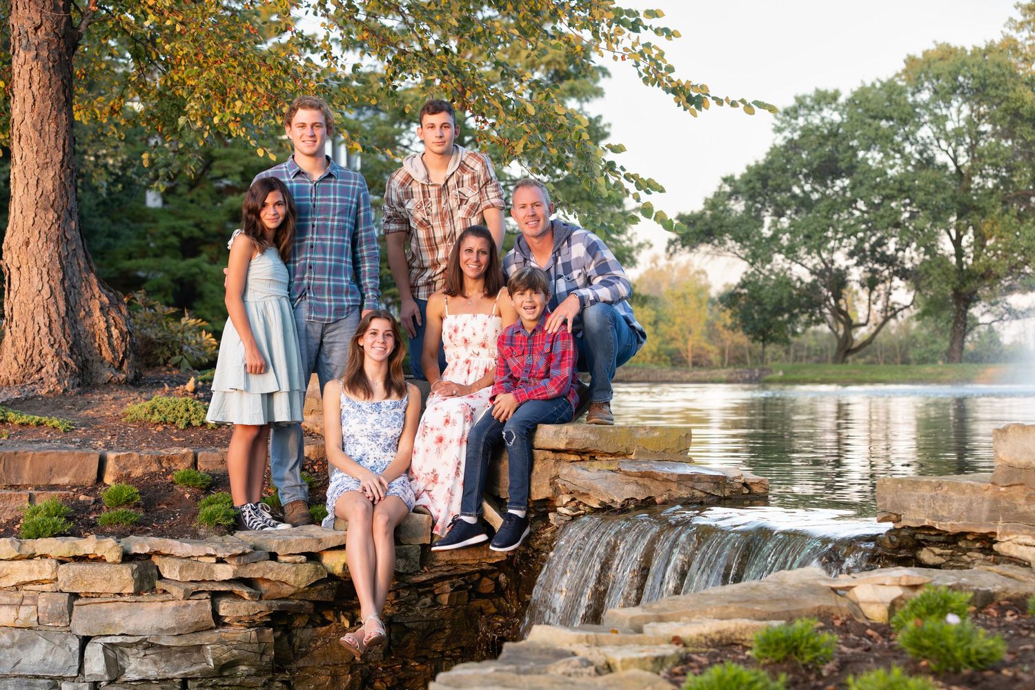 Multi-generational family posing together on stone waterfall steps near a scenic pond surrounded by lush green trees.