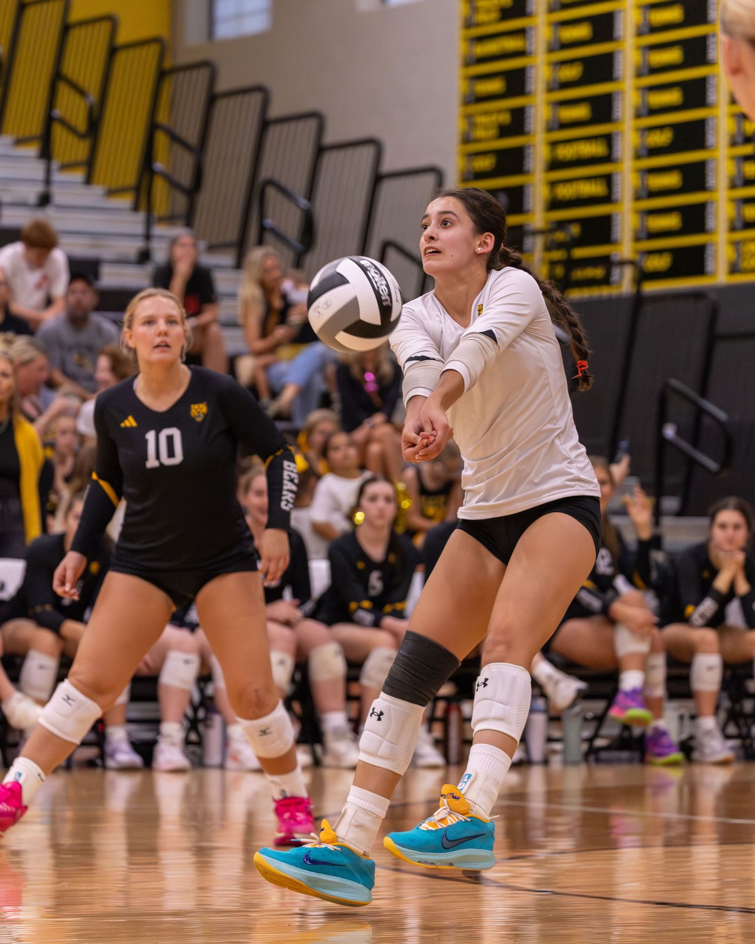 Female volleyball player in white uniform digs the ball during an indoor match while teammate in black watches.