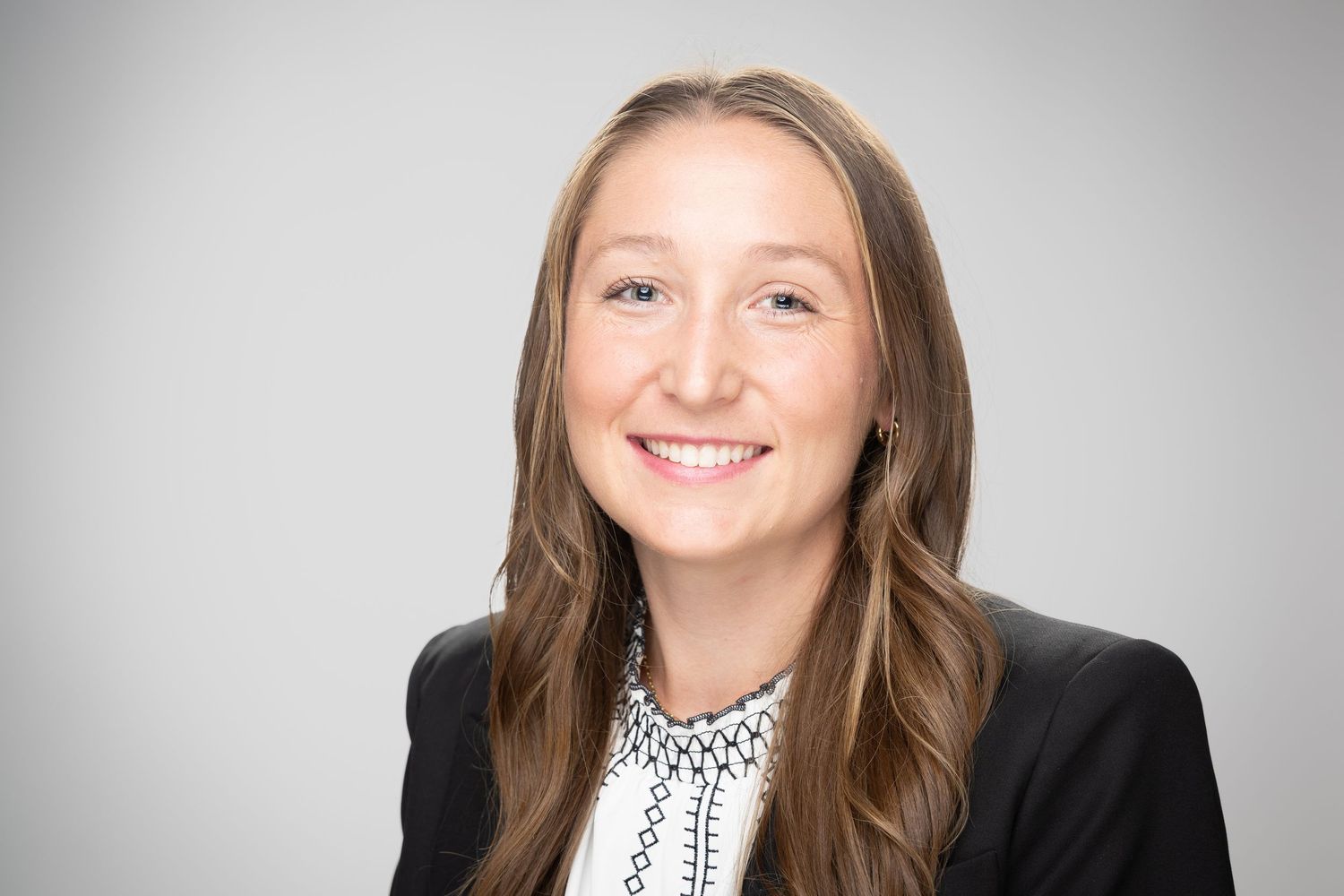 Professional headshot of a smiling young woman with long brown hair wearing a black blazer and white blouse.