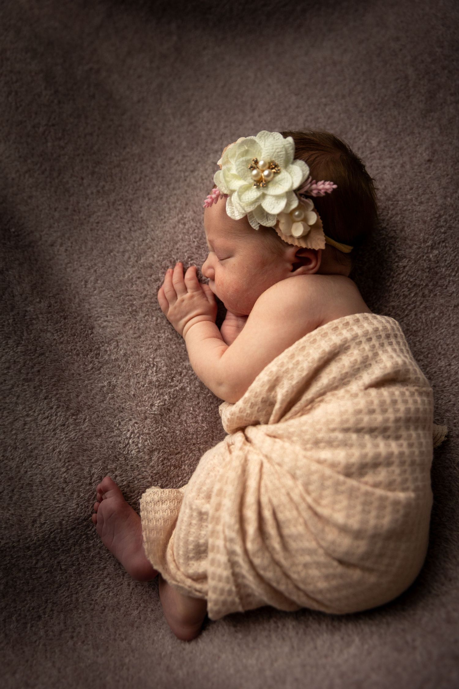 Sleeping newborn baby girl wrapped in cream knit blanket wearing floral headband on gray background.
