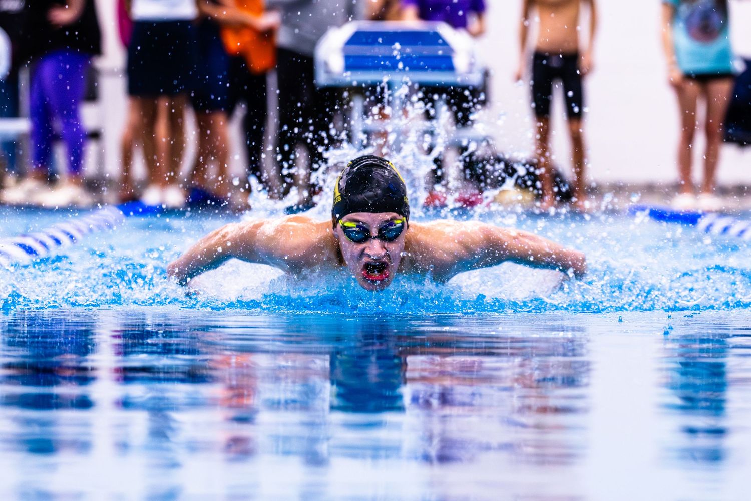 Competitive swimmer performing butterfly stroke in pool, wearing goggles and cap, with splashing water and spectators behind.