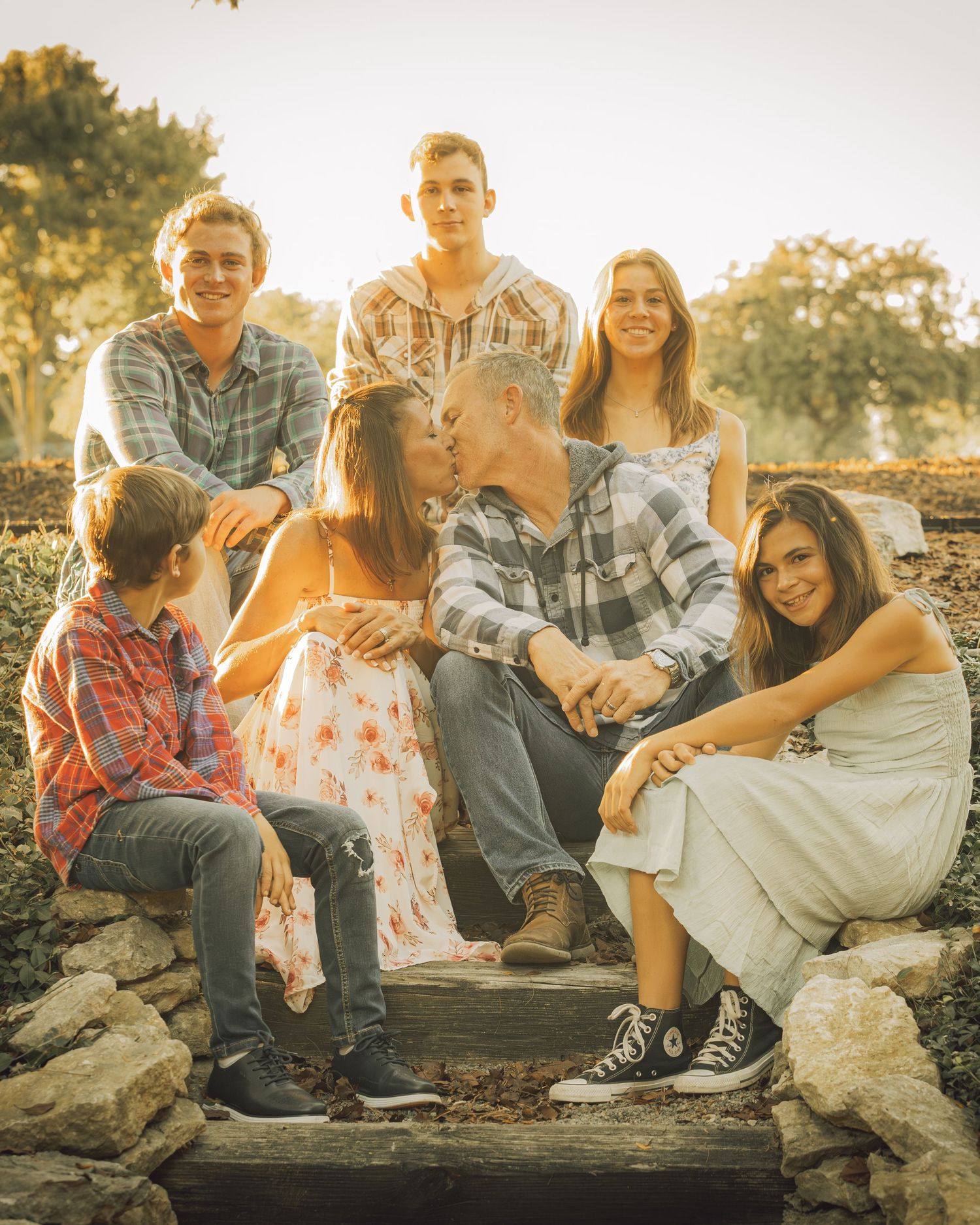 A family of seven poses outdoors at golden hour, with parents kissing in the center surrounded by their smiling children.