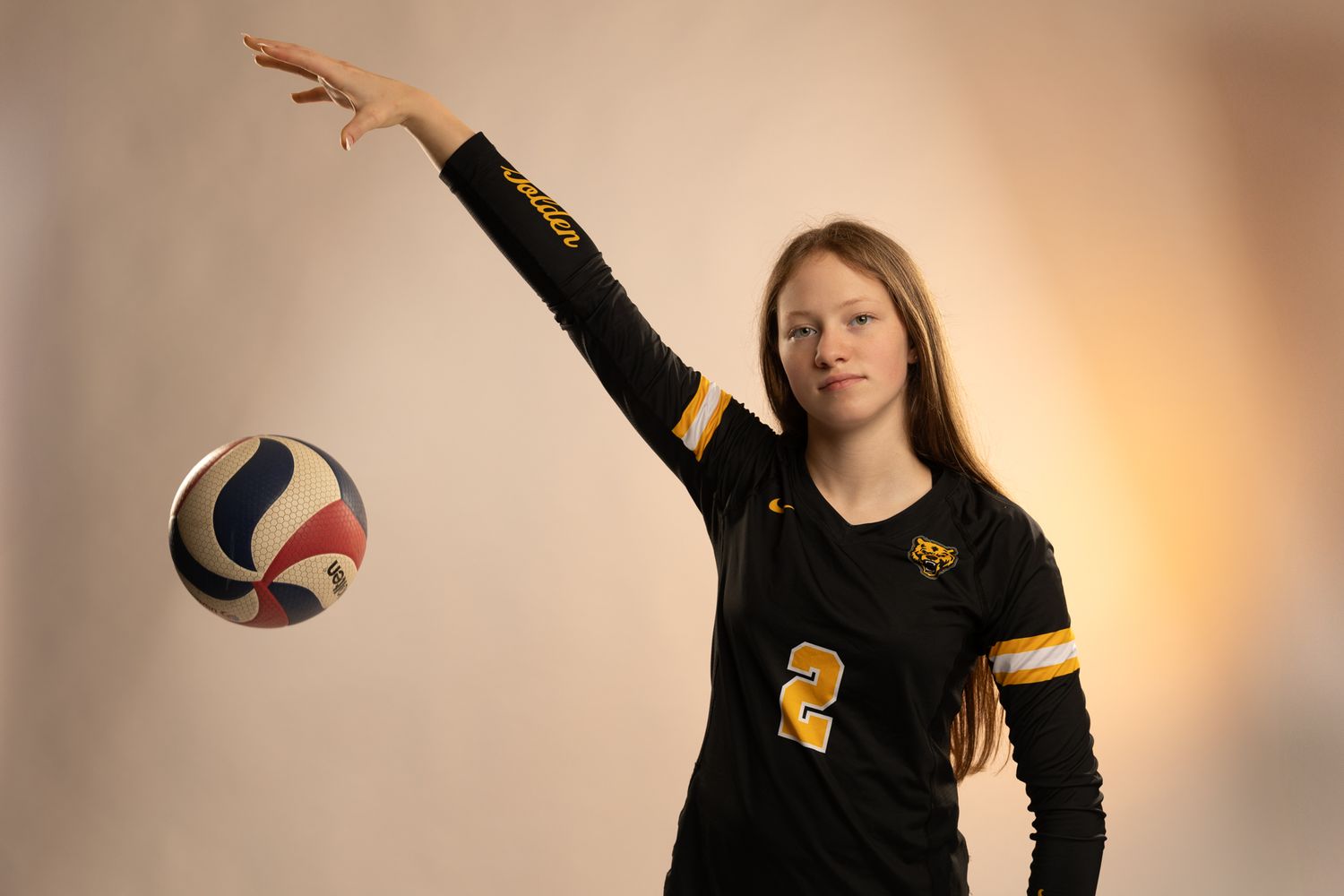 Young female volleyball player in black and gold uniform, number 2, posing with ball and raised arm.