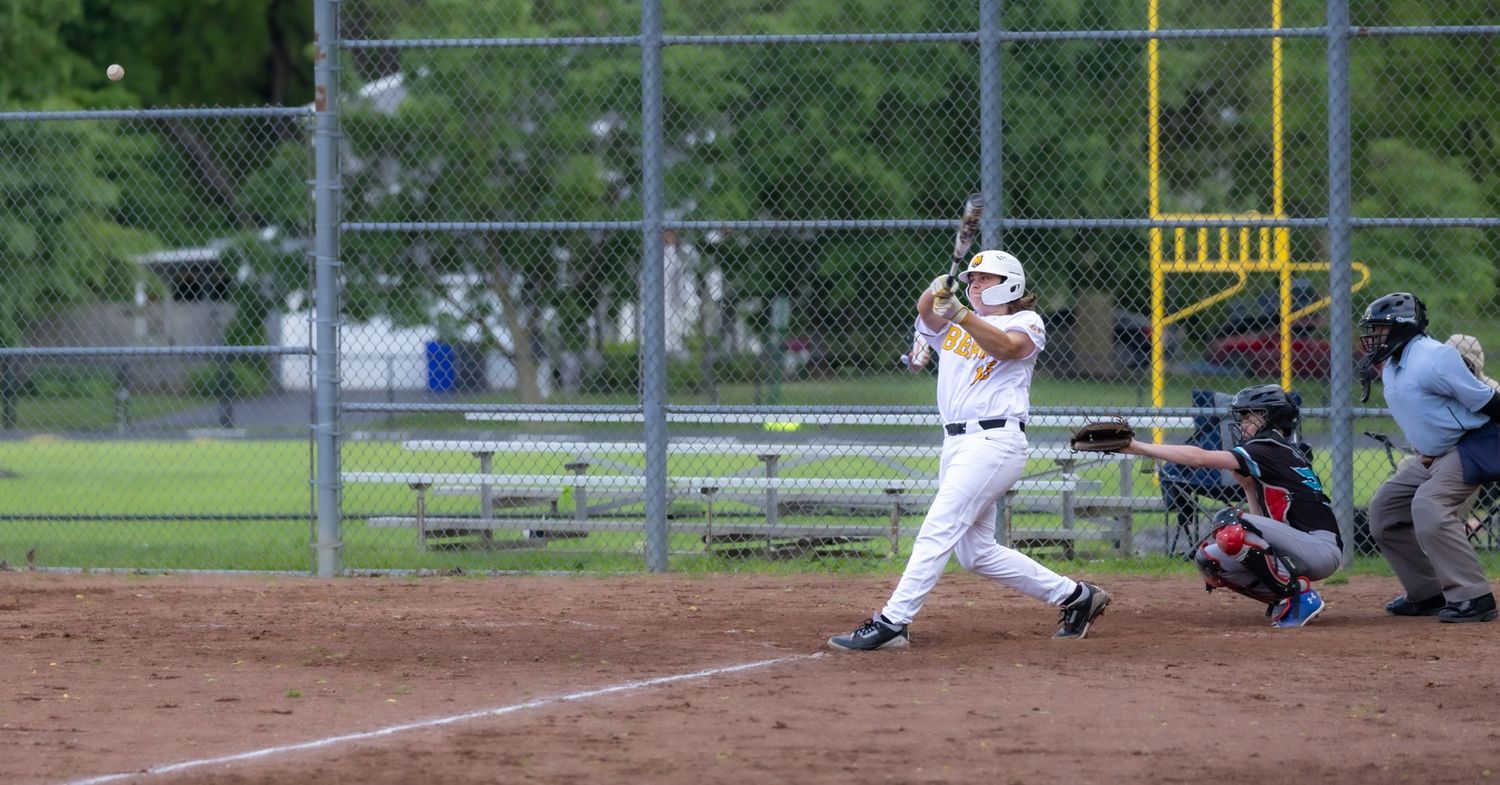 Baseball batter in white uniform swinging at pitch during youth league game with catcher and umpire behind home plate.