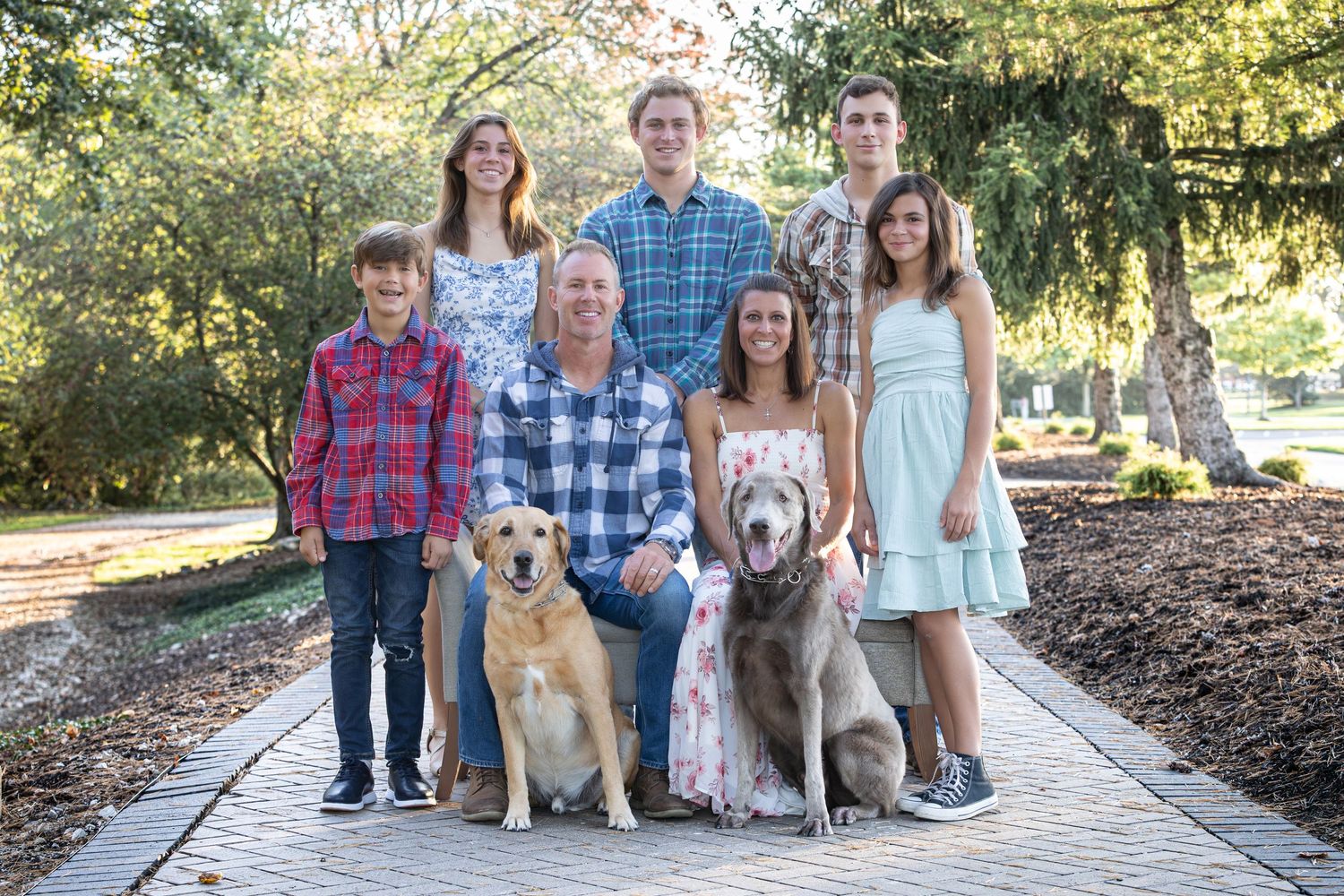 Family of six poses with two dogs on a brick path in a lush green park setting for a portrait.