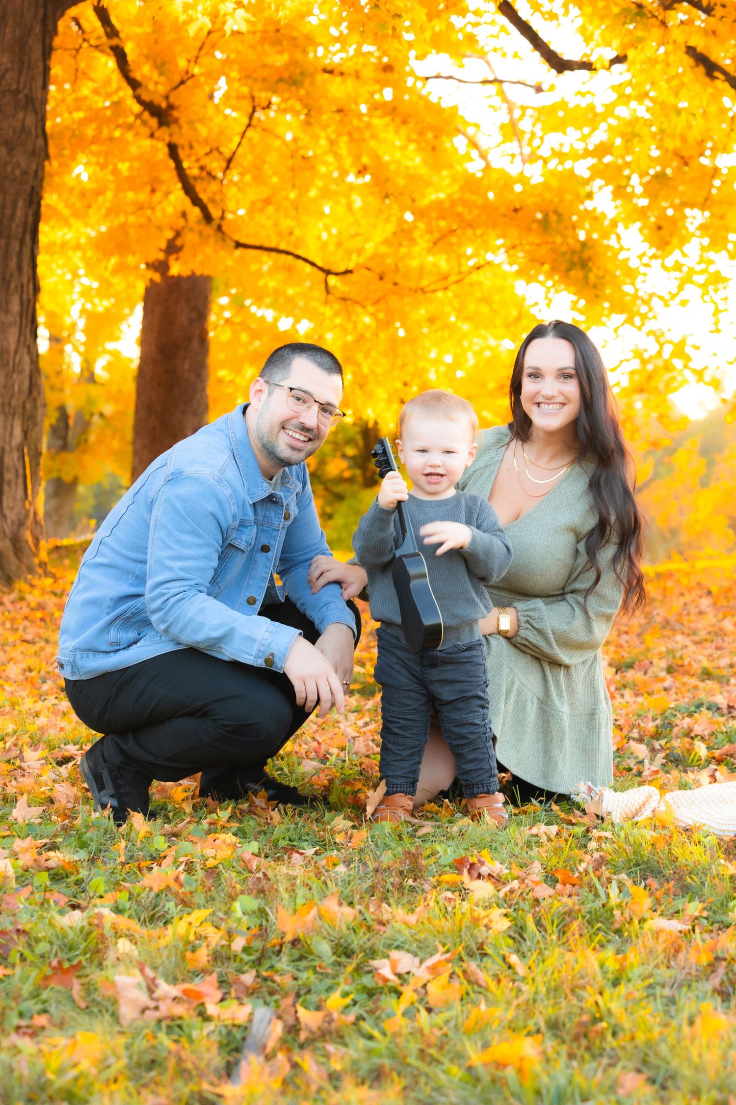 Family of three posing outdoors in autumn park with vibrant yellow foliage, parents smiling with toddler standing between them.
