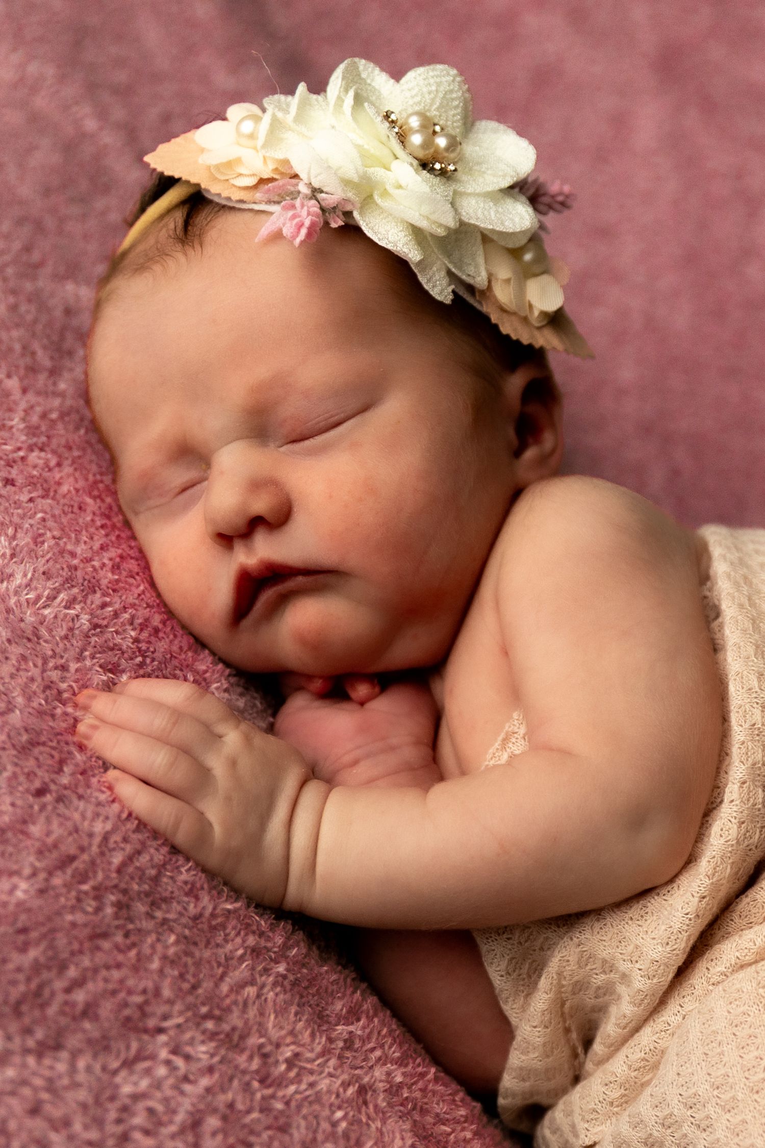 Sleeping newborn baby girl wearing a delicate floral headband, wrapped in a soft blanket on a pink background.