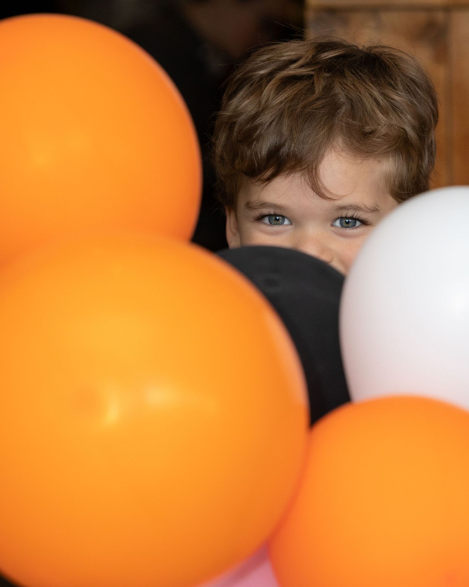A young child peers curiously through a cluster of orange, black, and white Halloween balloons.