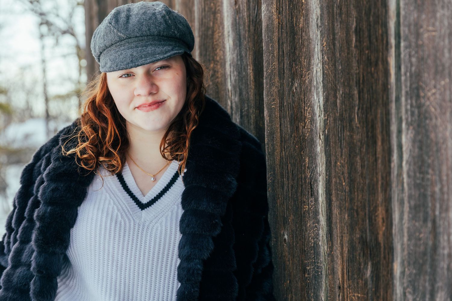 Young woman with red hair wearing a gray newsboy cap and black fur coat leaning against a rustic wooden wall.