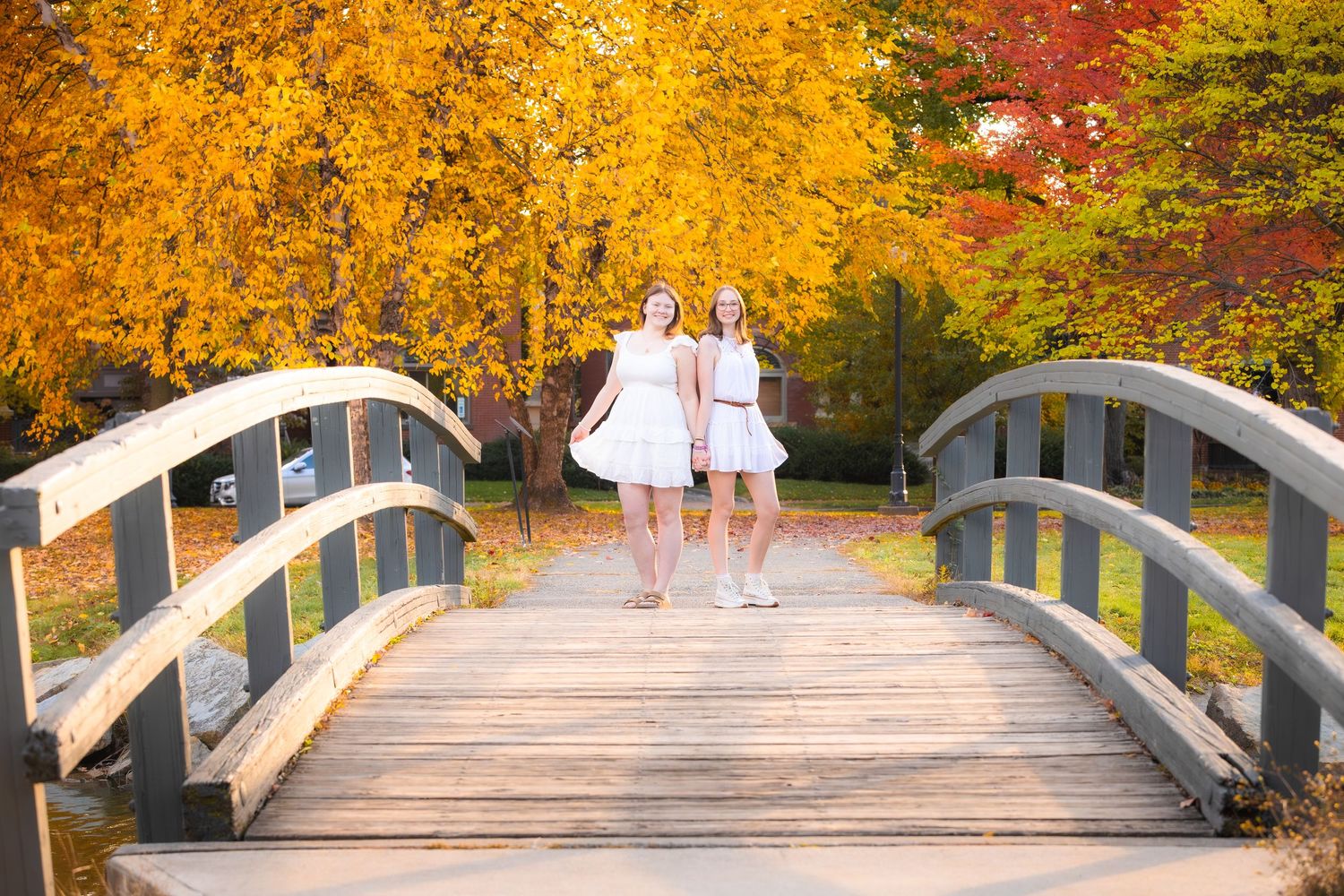 Two girls in white dresses walk on a wooden bridge surrounded by vibrant autumn foliage in a park.
