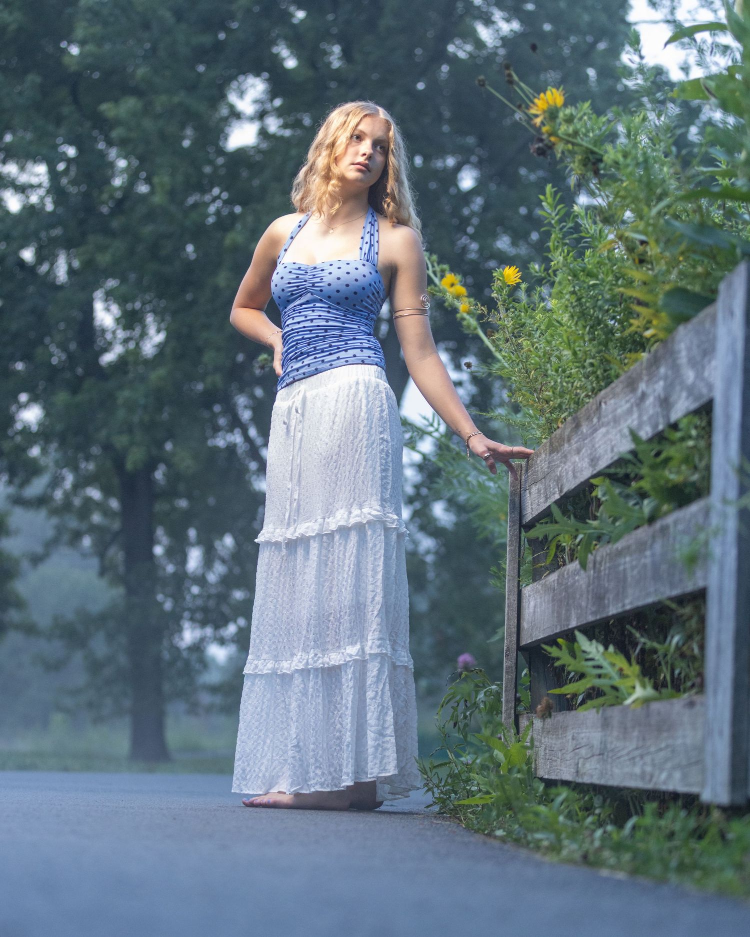 Young blonde woman in blue smocked top and white tiered maxi skirt stands by rustic wooden fence on misty rural road.