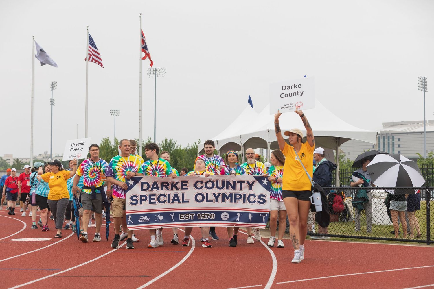 Darke County Special Olympics athletes march on track with banner during opening ceremony with flags in background.