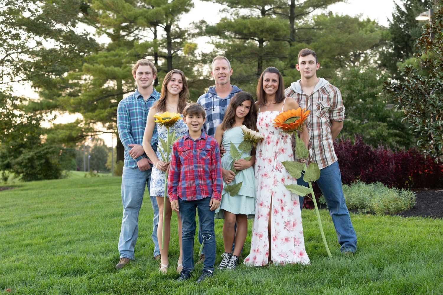 A cheerful family of seven posing outdoors on green grass, holding sunflowers, dressed in casual attire with pine trees behind.