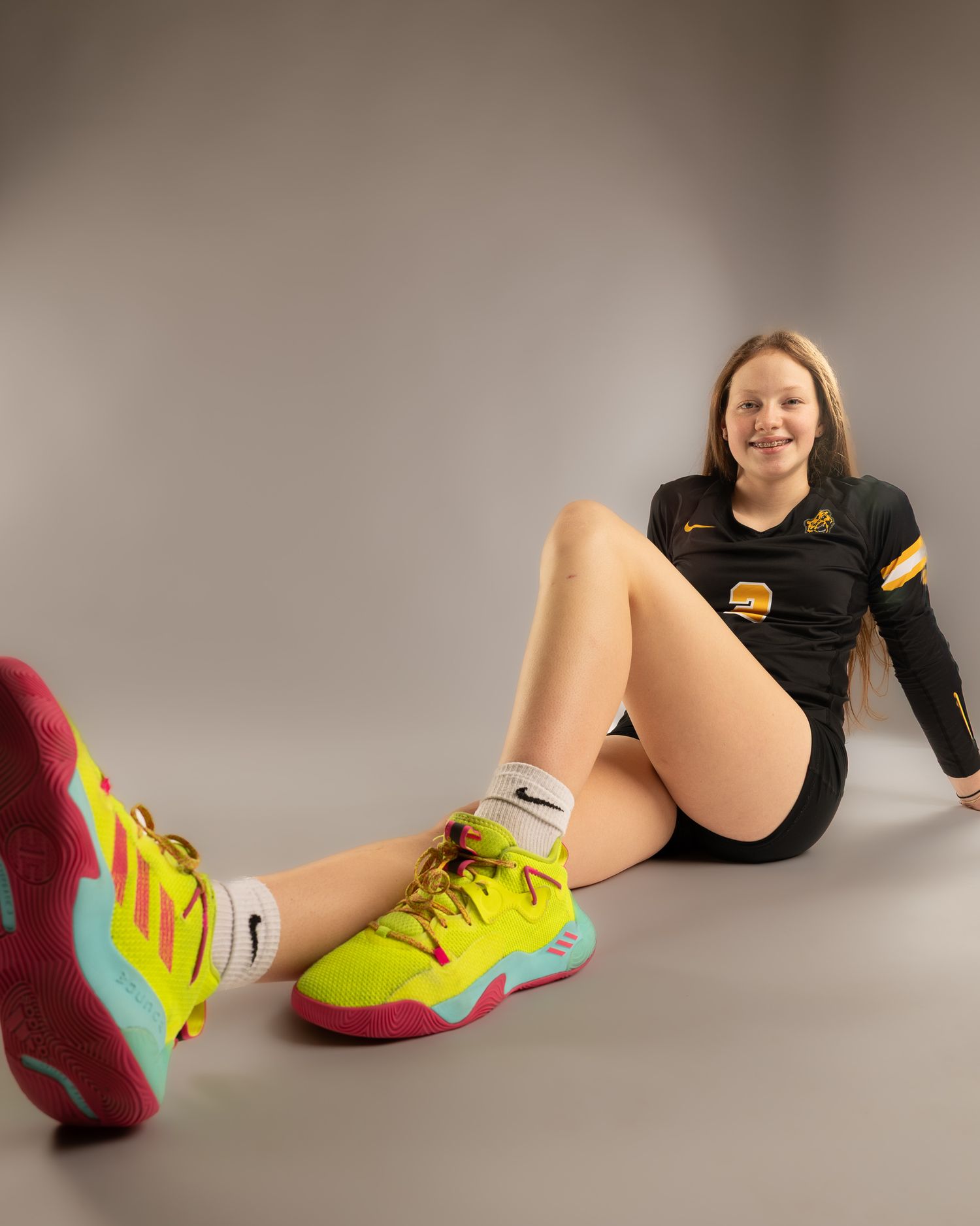 Young female volleyball player in black Nike uniform and bright yellow Nike shoes poses seated on gray studio floor.