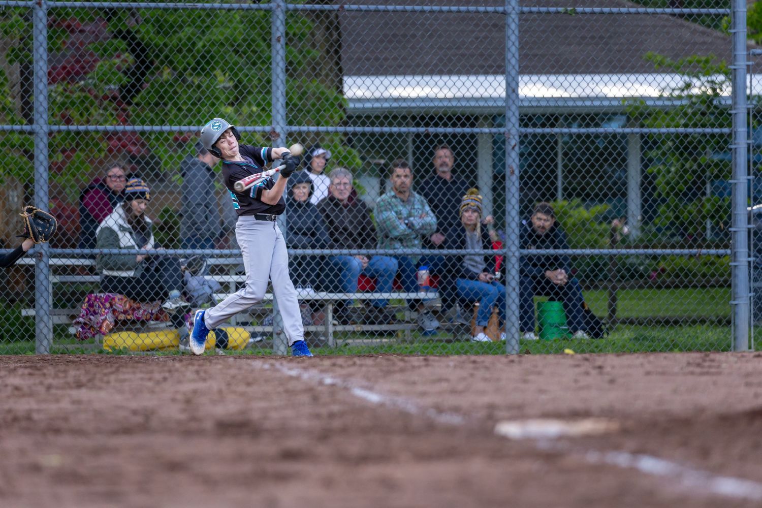 Youth baseball player swings bat during a game with spectators watching from bleachers behind a chain-link fence.