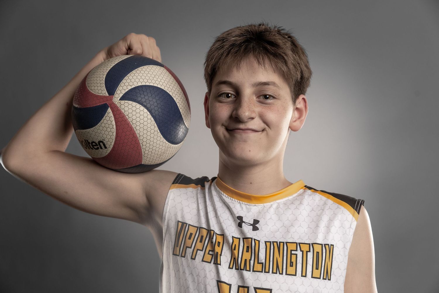 Young athlete in Upper Arlington jersey holding a volleyball, smiling confidently against a gray background.