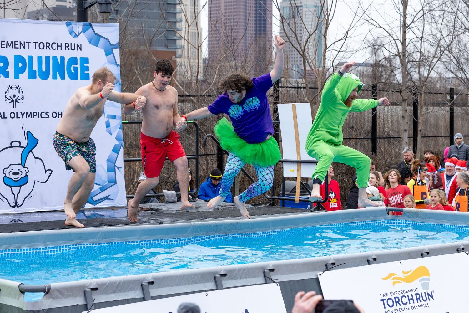 Participants in costumes leap into a pool during the Special Olympics Polar Plunge fundraiser event outdoors.