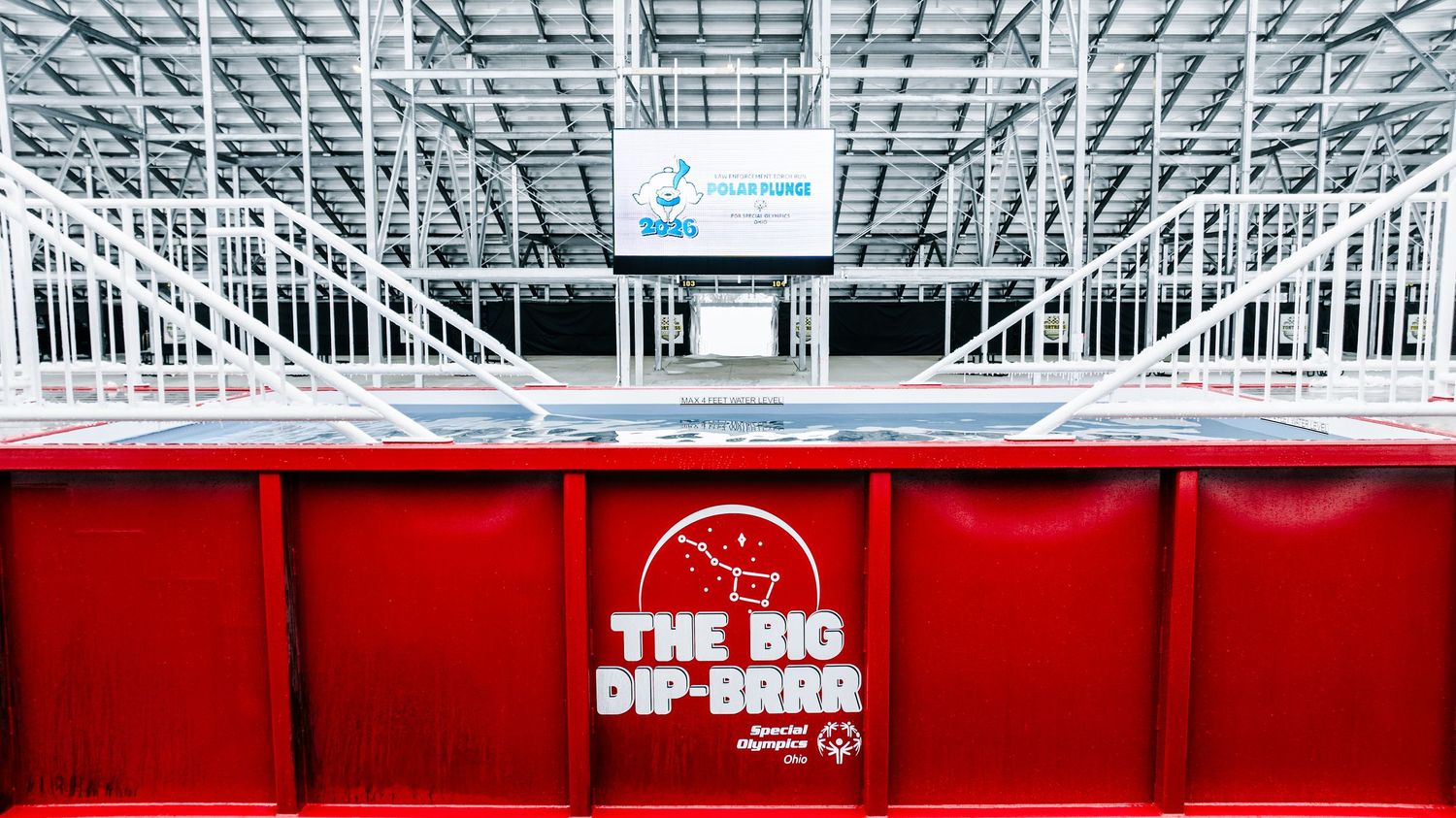 Red padded barrier with The Big Dip-Brah logo inside a large indoor ice skating rink with white steel framework.
