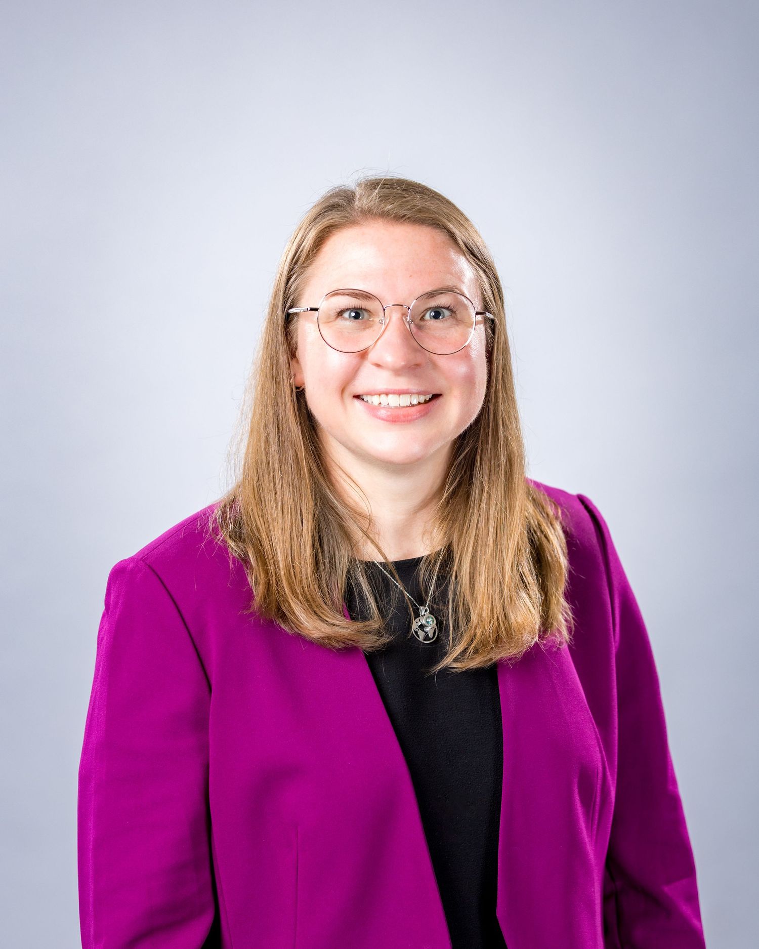 Professional headshot of someone wearing glasses and a magenta blazer with a black top against a light background.