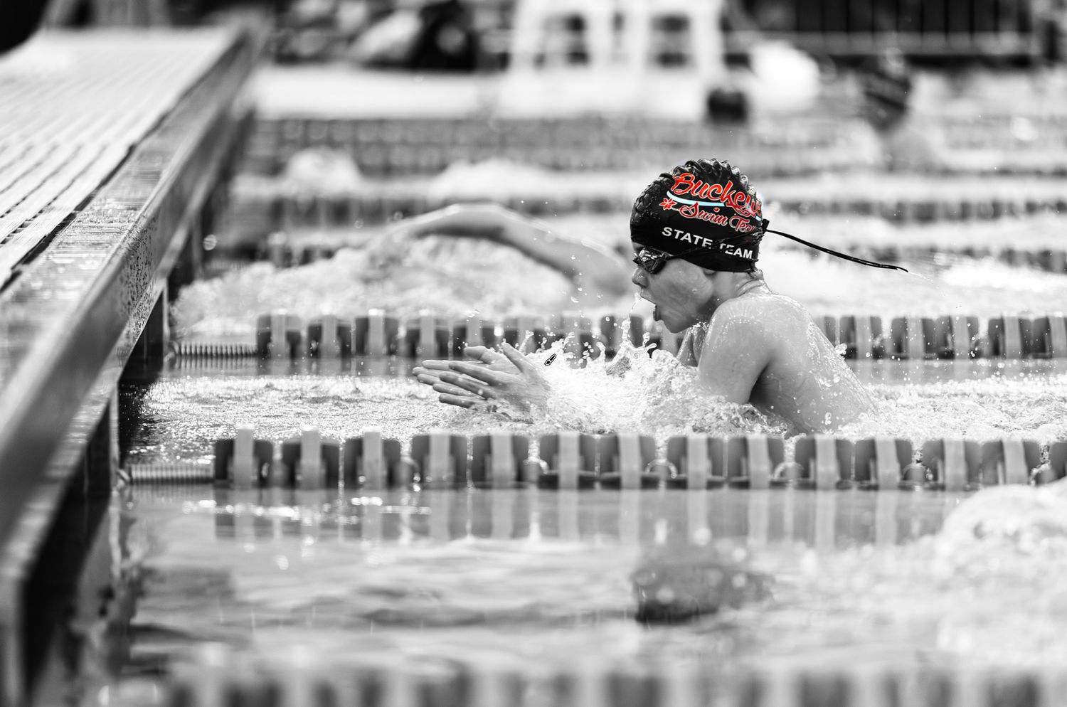 Swimmer in red cap competes in breaststroke at a swim meet, captured in dramatic black and white photography.