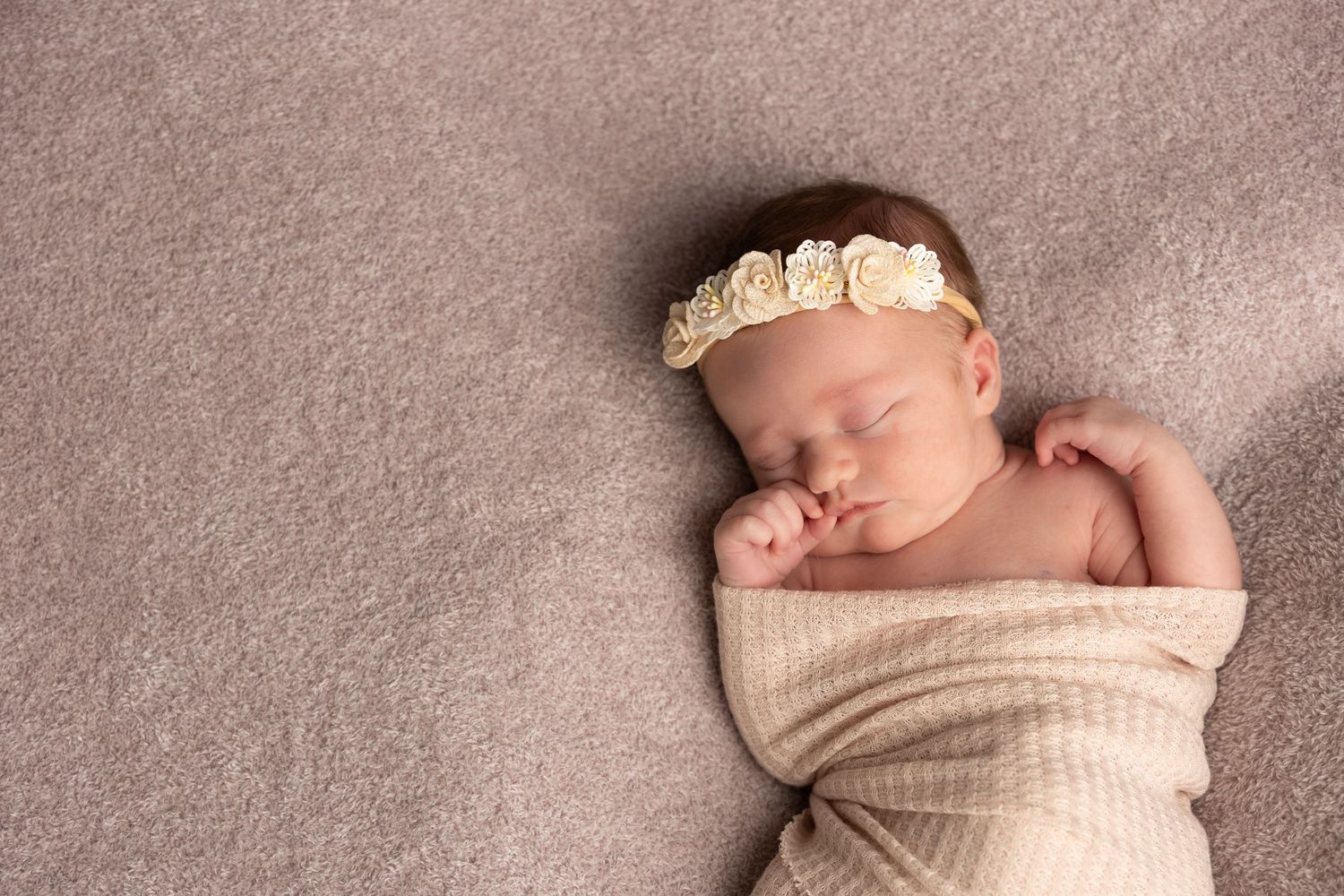 Peaceful newborn baby girl sleeping swaddled in cream wrap with floral headband on soft taupe background.