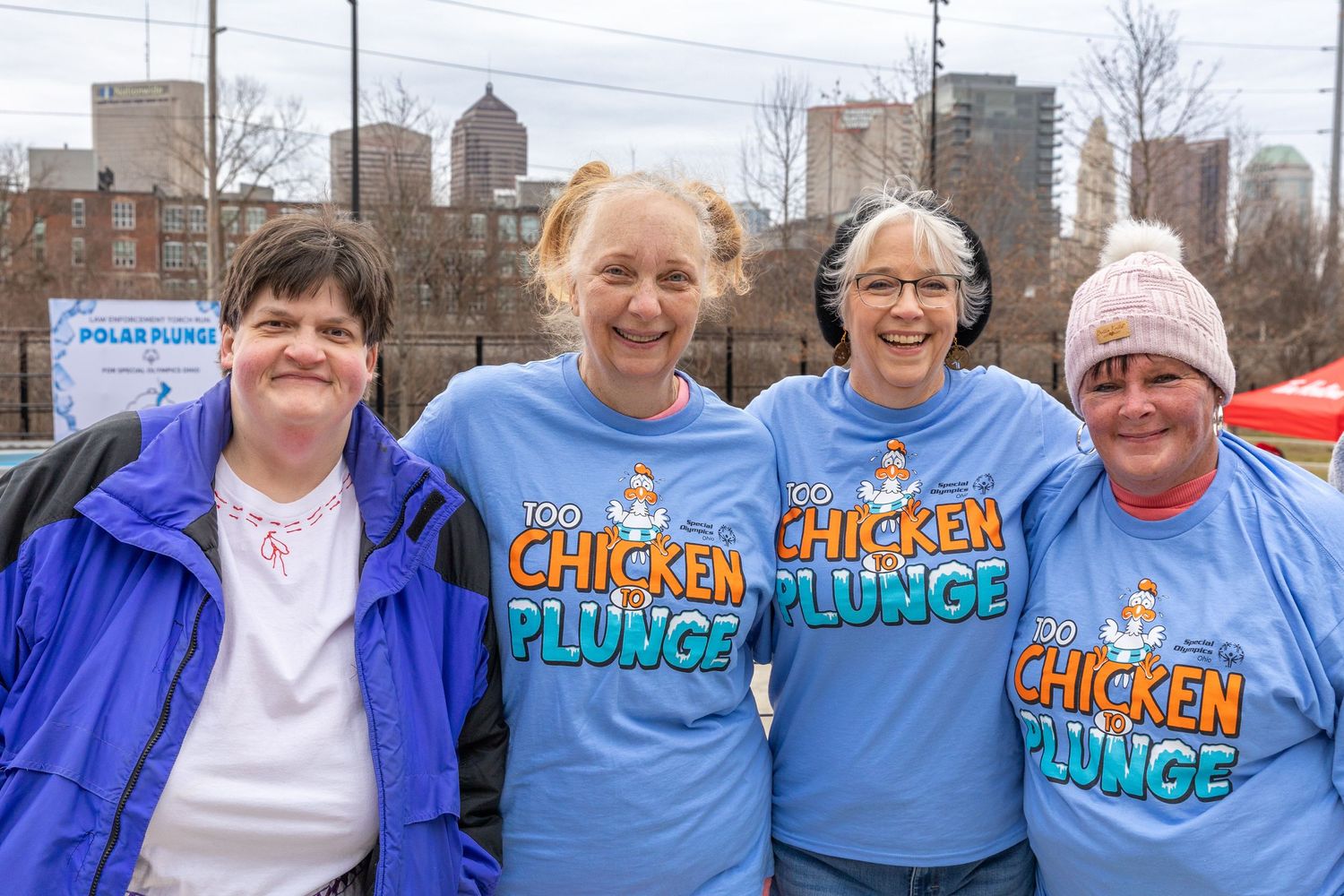 Four women smiling outdoors wearing blue Chicken Plunge charity event t-shirts at a Polar Plunge fundraiser.