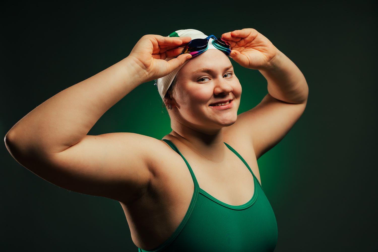 Female swimmer in green swimsuit adjusting goggles on swim cap, smiling confidently against dark background.