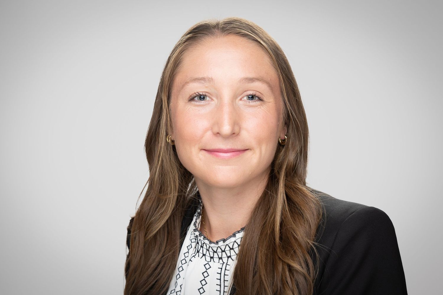 Professional headshot of a smiling woman with long brown hair wearing a black blazer and white patterned blouse.