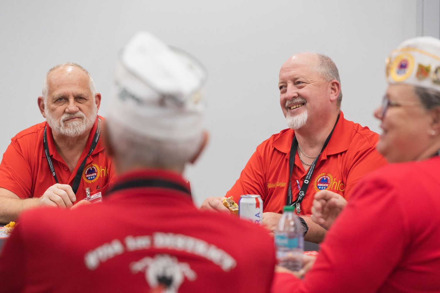 Veterans in red polo shirts share laughs at a table during a meeting, with some wearing white military caps.