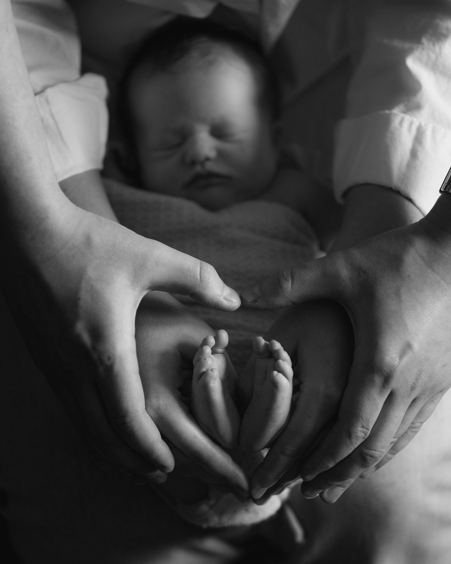 Black and white photo of sleeping newborn baby cradled in parents hands forming a heart shape around tiny feet.