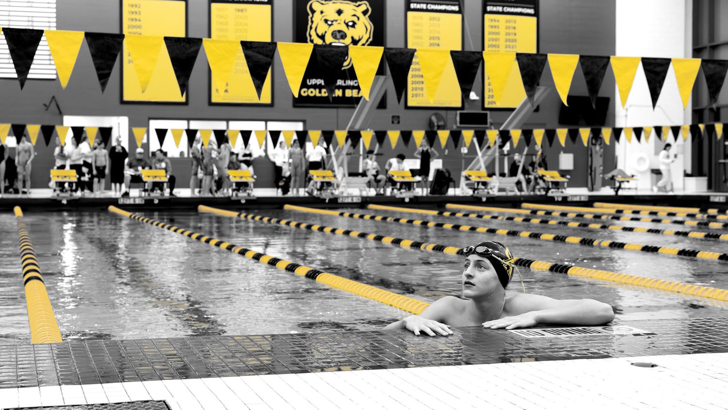 Swimmer competes in indoor pool at a black and gold themed swim meet with lane dividers and spectators.