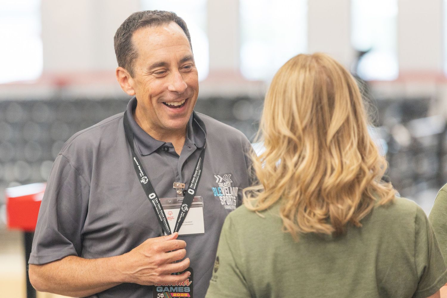 A smiling man wearing a lanyard converses with a blonde woman indoors at a casual event.